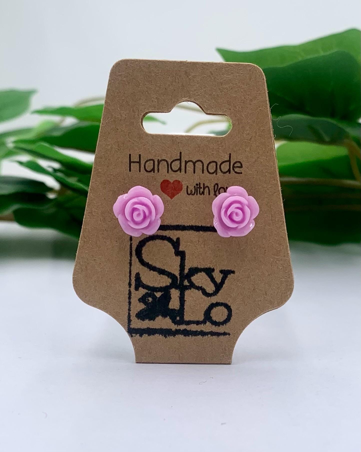 Pair of lavender rose-shaped earrings on a 'Handmade with love' branded card with a blurred green leafy background.