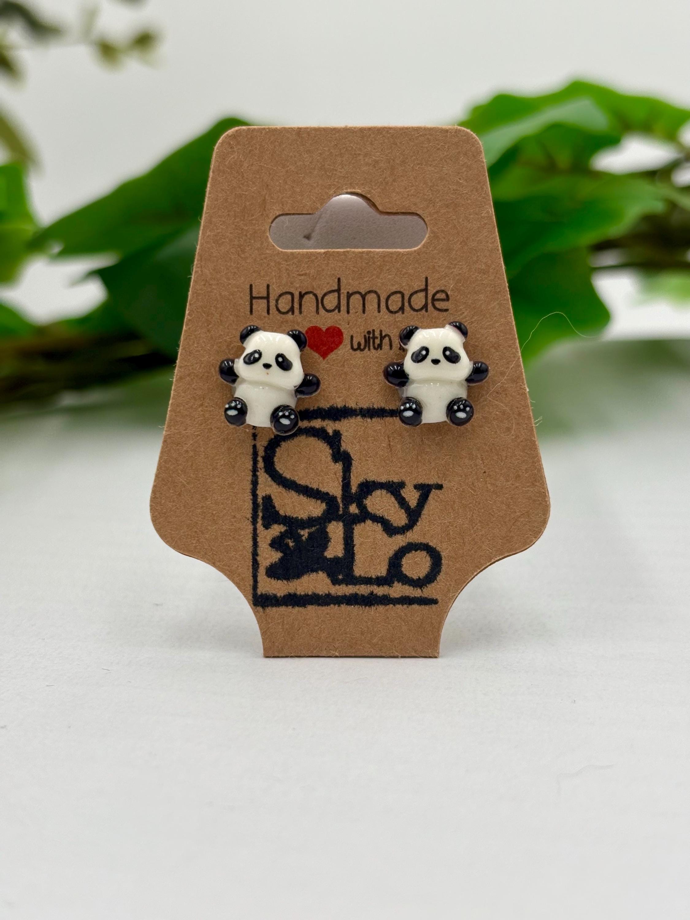 Panda-shaped earrings on a brown card with 'Handmade with love' branding, against a green leafy background.