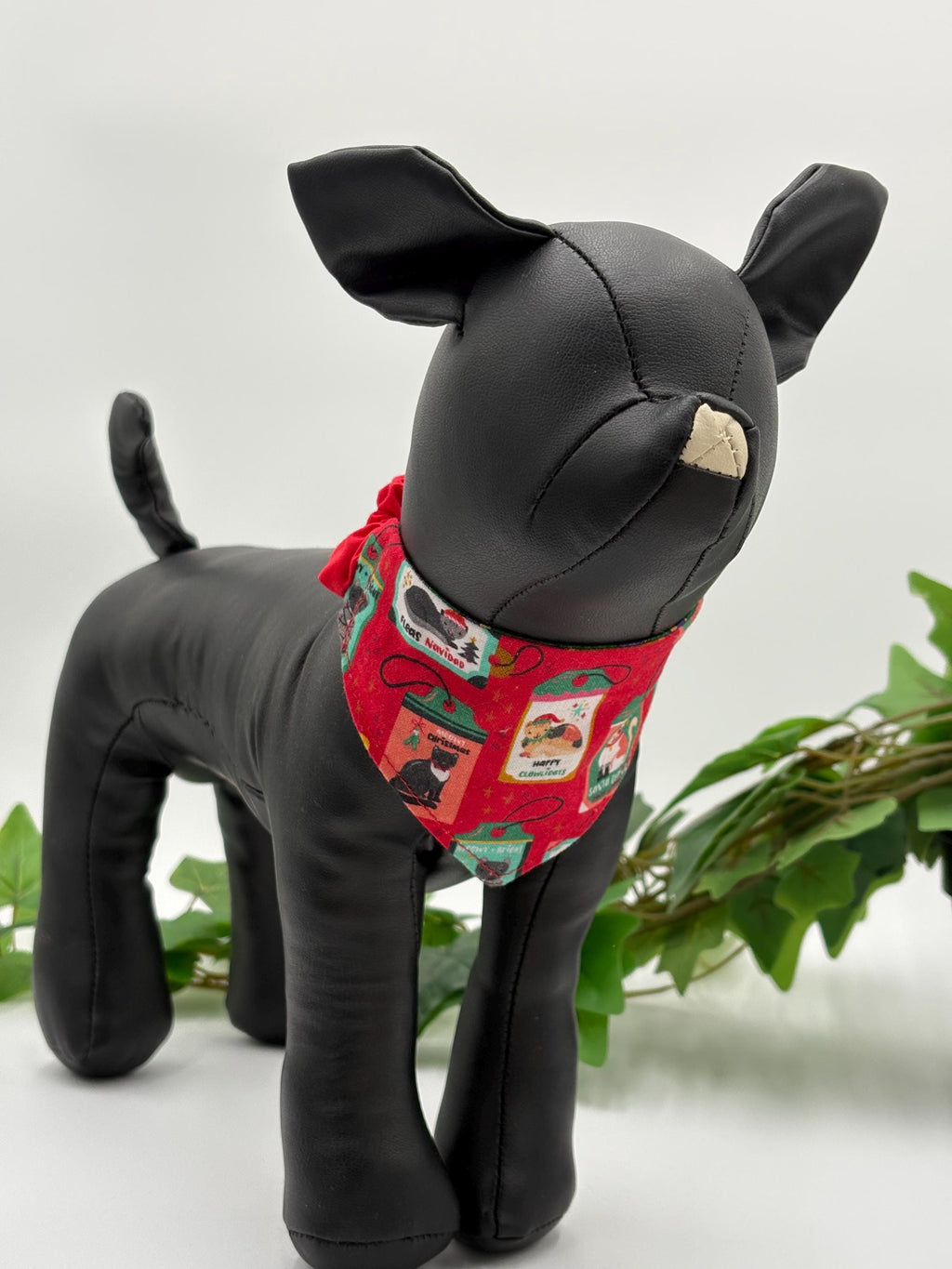 Black dog mannequin with a red bandana featuring cats on a white background standing next to a potted plant. 