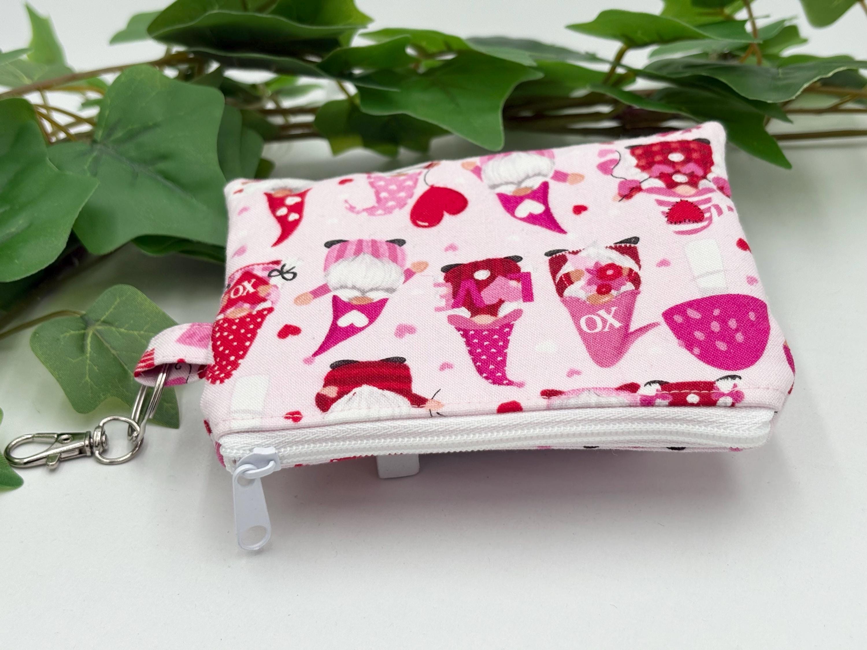 Small coin purse keychain with Valentine-themed gnomes and hearts pattern with white zipper on a white background with green leaves