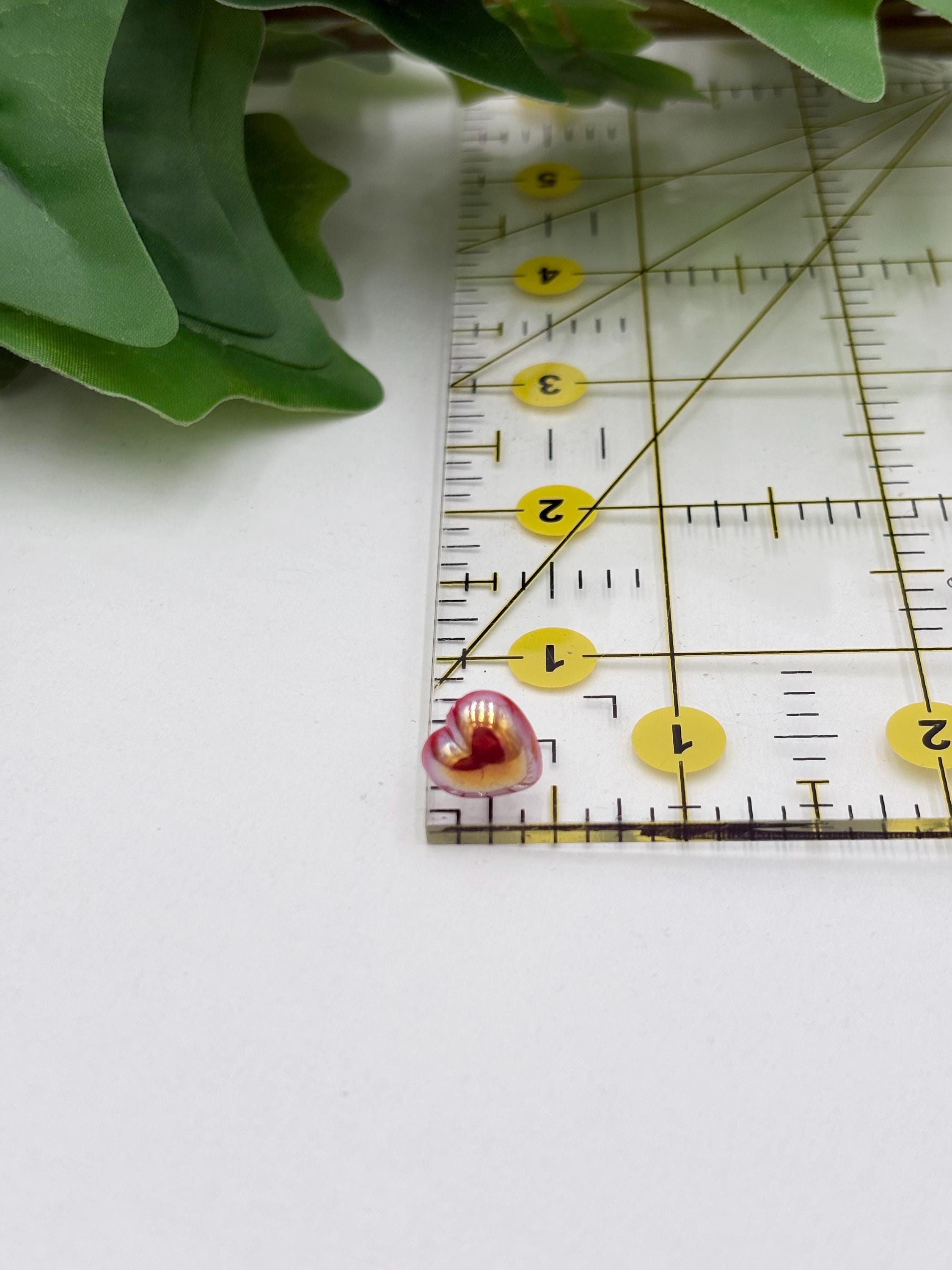 Small pink metallic heart shaped earring next to a clear ruler with yellow markings on a white background