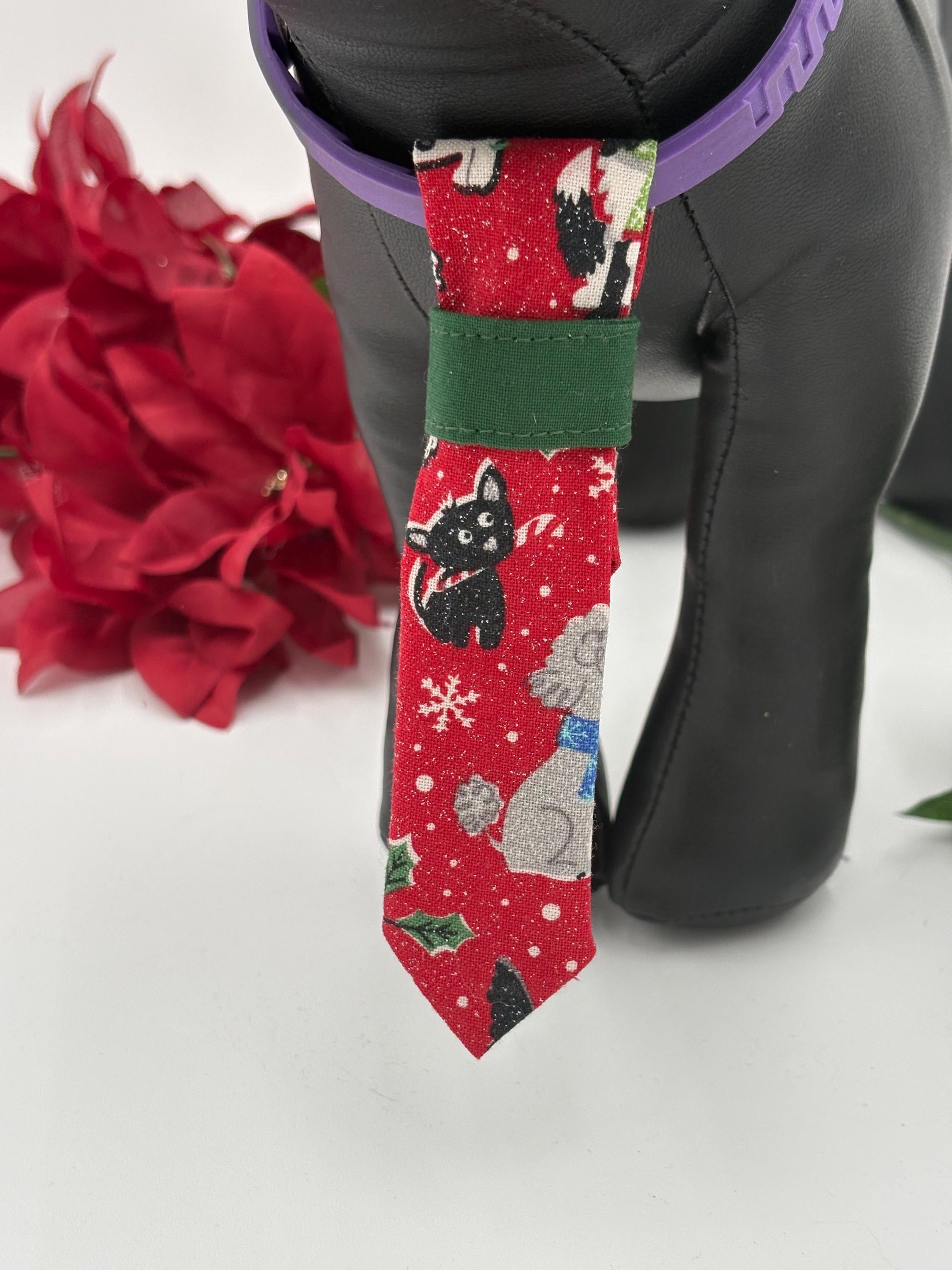 close up of Black dog mannequin wearing a red over the collar necktie with a dog christmas pattern, surrounded by red flowers and greenery on a white background.