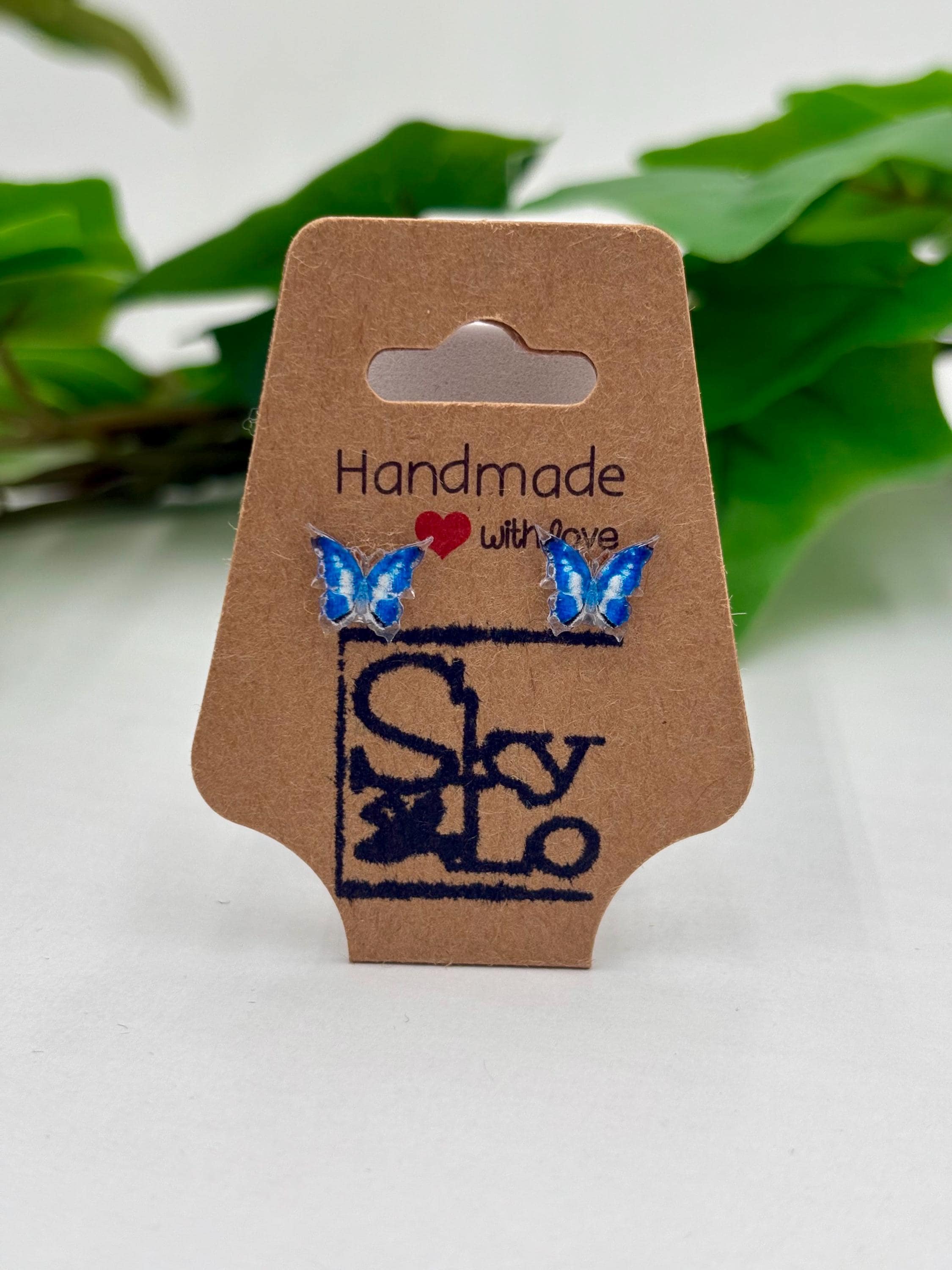 Blue Butterfly earrings on a 'Handmade with Love' branded card with a blurred green leaf background.