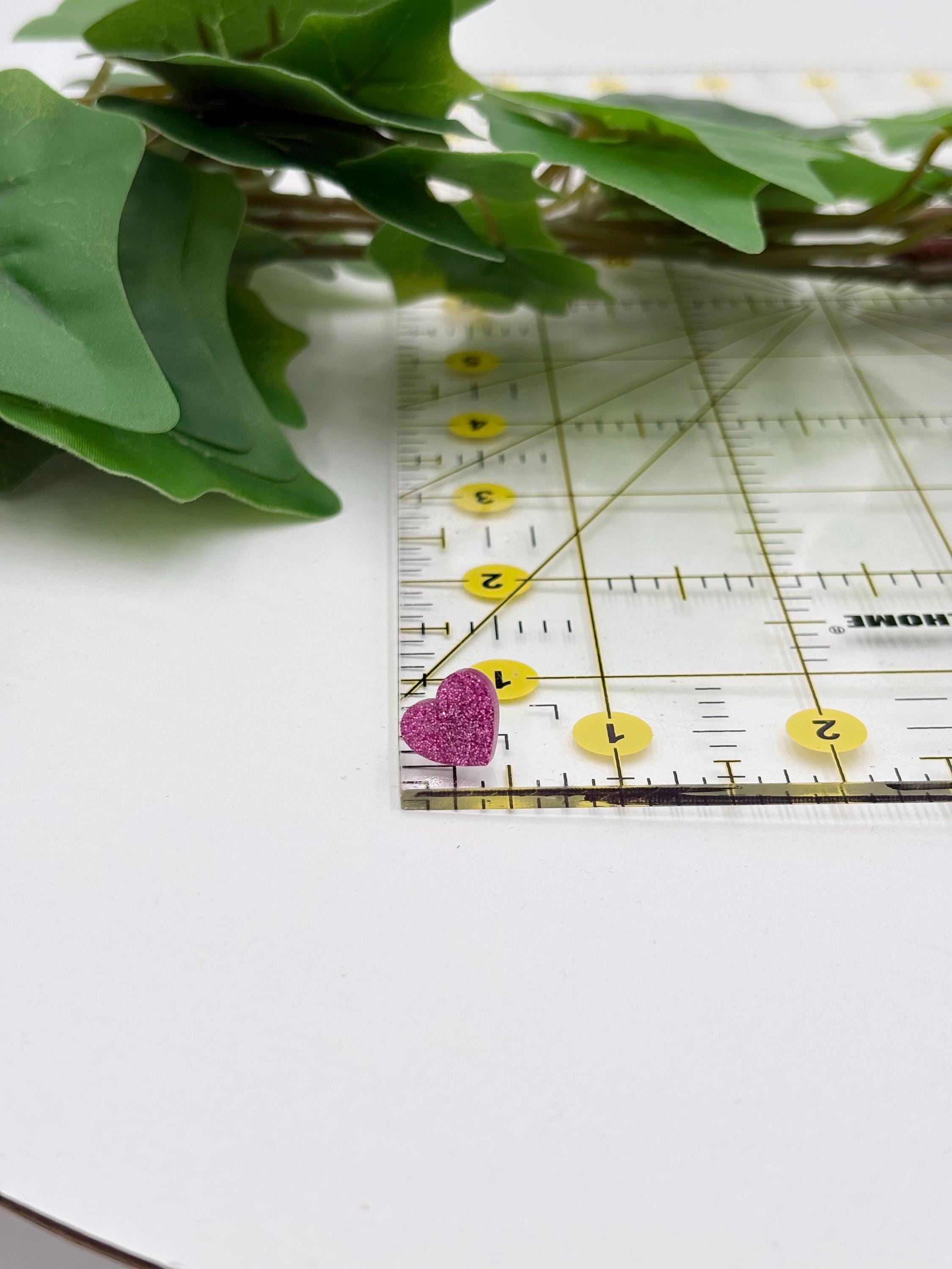 Ruler with pink sparkling heart-shaped earring on a white surface with green leaves.