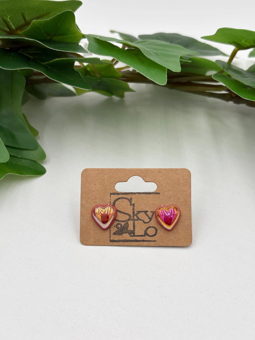 Pink metallic Heart-shaped earrings on display with a brand label against a white background with green leaves.