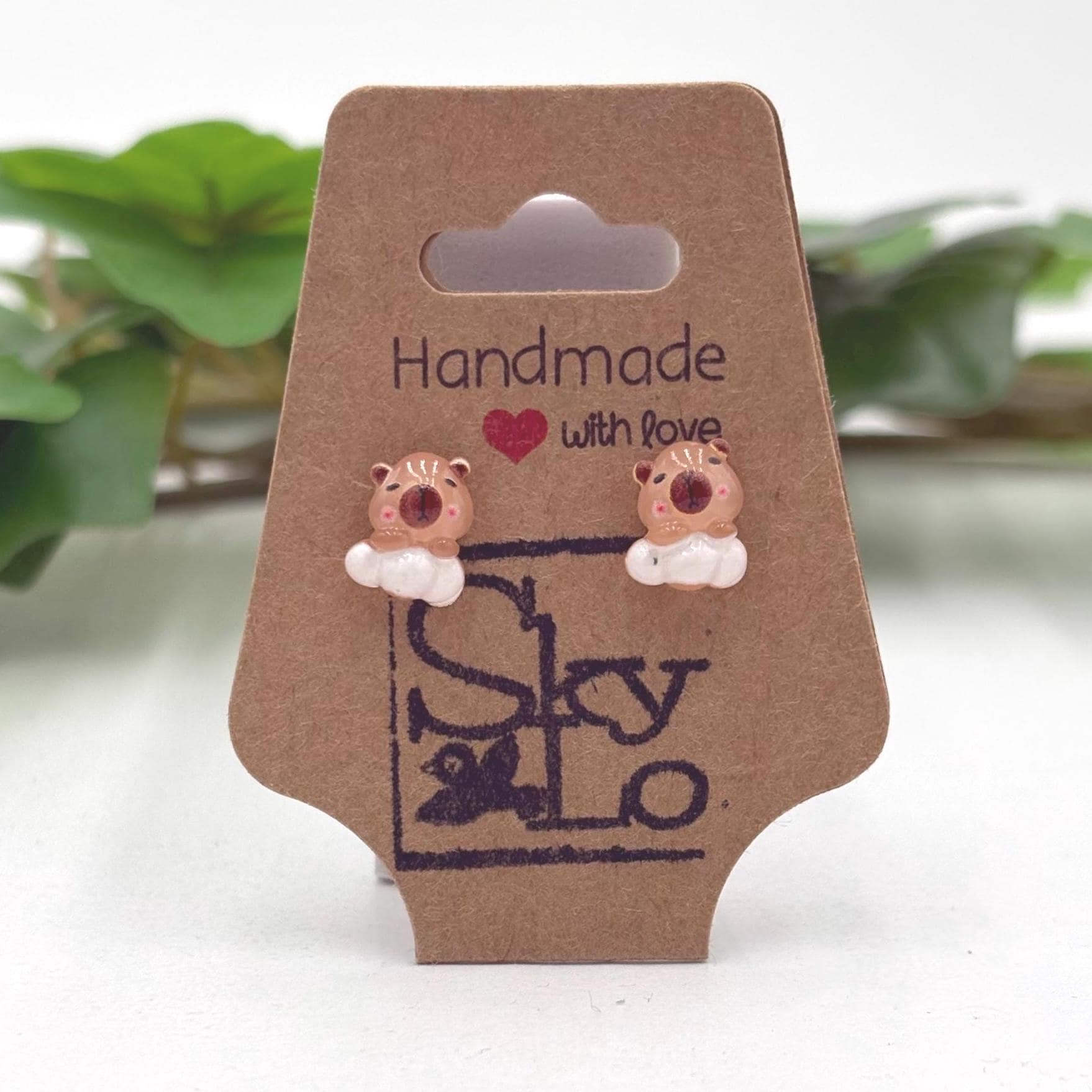 Handmade capybara on a white cloud earrings on a branded card with a blurred green plant background