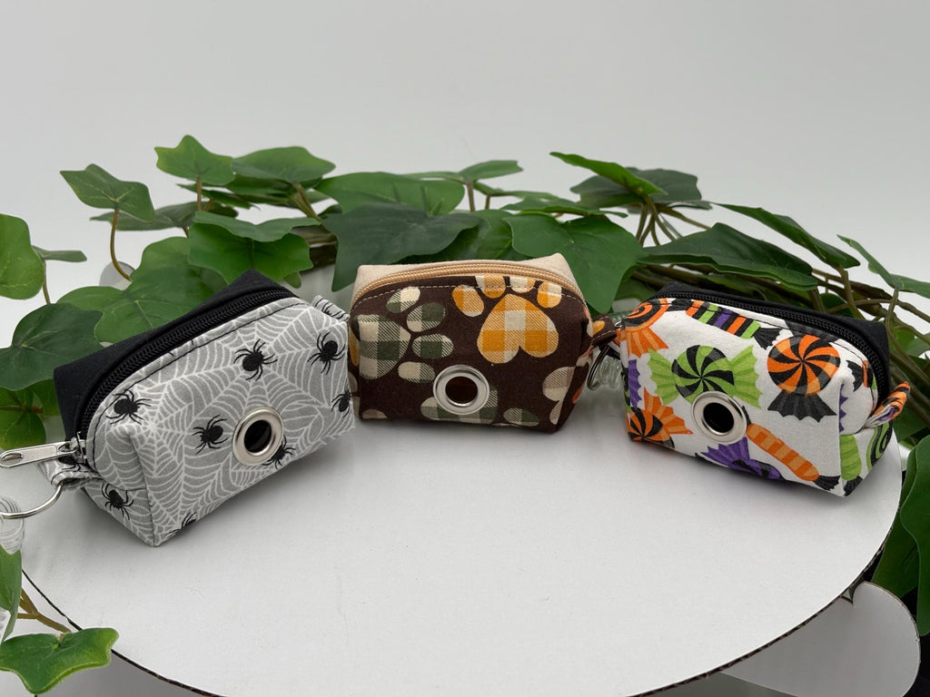 Three Autumn and Halloween themed dog poop bag holders on a white surface with green leaves in the background