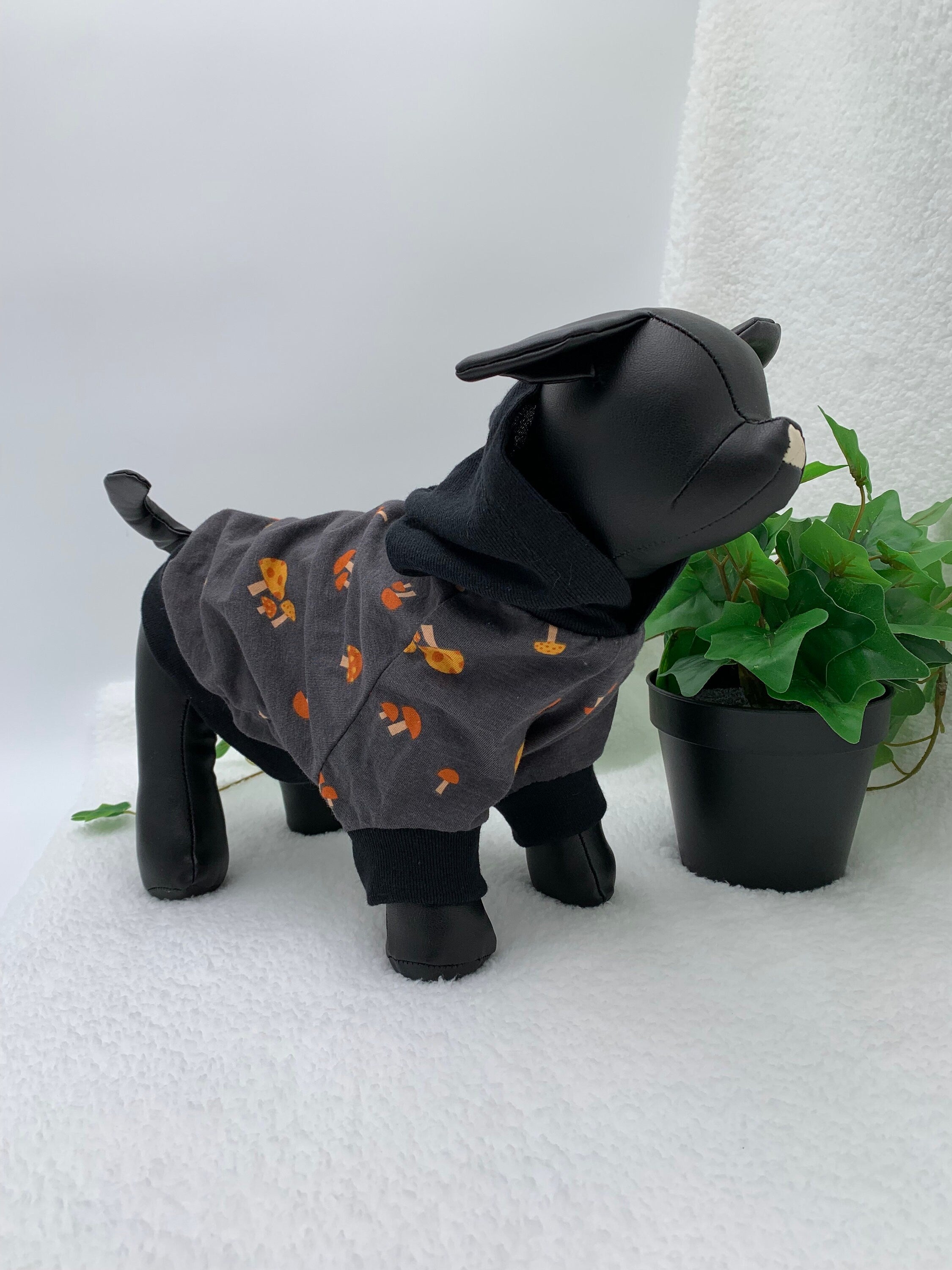 Black dog mannequin wearing a gray mushroom patterned hoodie with hoodie down, standing next to a potted plant on a white surface.