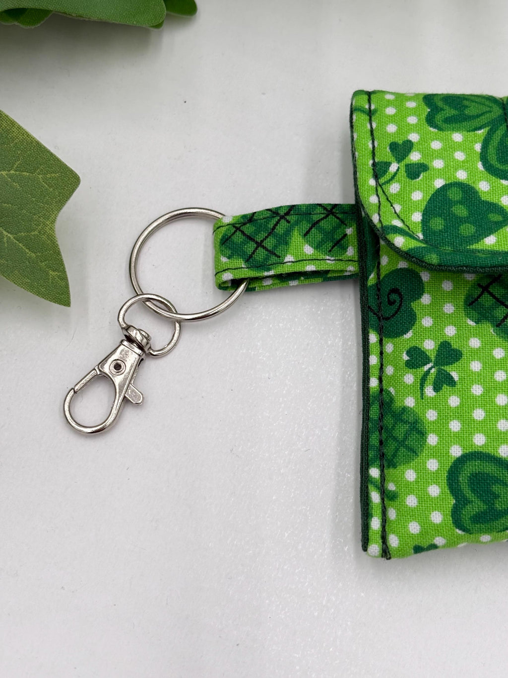 Close up of lobster clasp keychain for a Small quilted headphone holder pouch keychain with green shamrocks design on a white surface with green leaves.