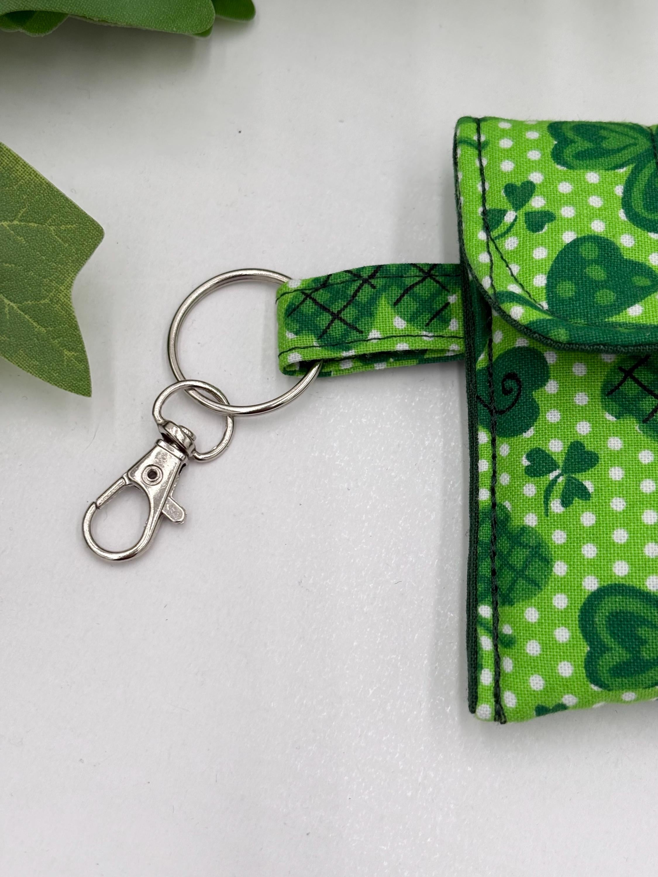 Close up of lobster clasp keychain for a Small quilted headphone holder pouch keychain with green shamrocks design on a white surface with green leaves.