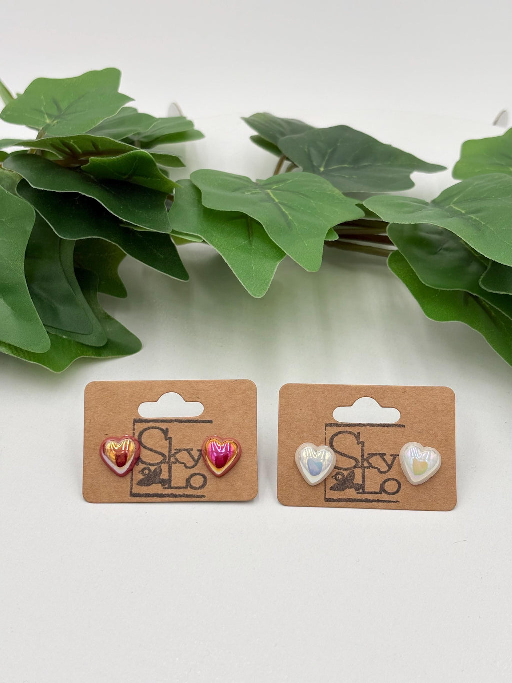 Two sets of metallic Heart-shaped earrings on display with a brand label against a white background with green leaves.