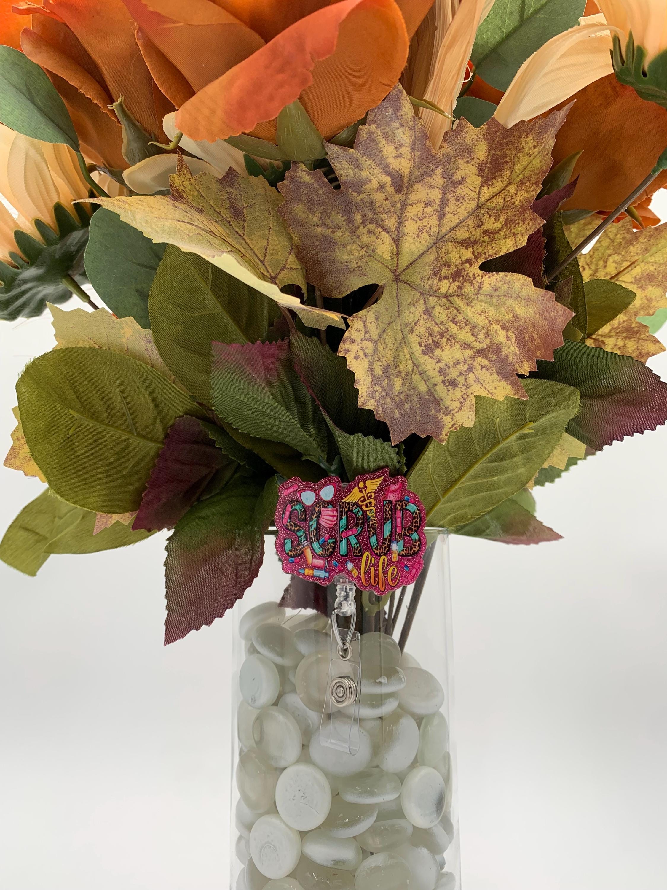 Bouquet of autumn leaves and flowers in a clear vase with decorative elements and a badge reel that says "Scrub Life"