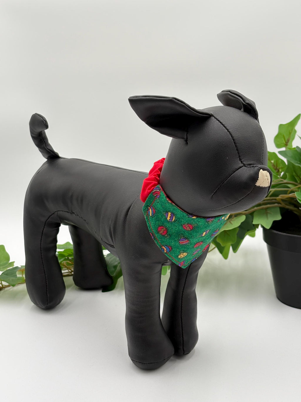 Black dog mannequin with a green bandana featuring mini ornaments on a white background standing next to a potted plant