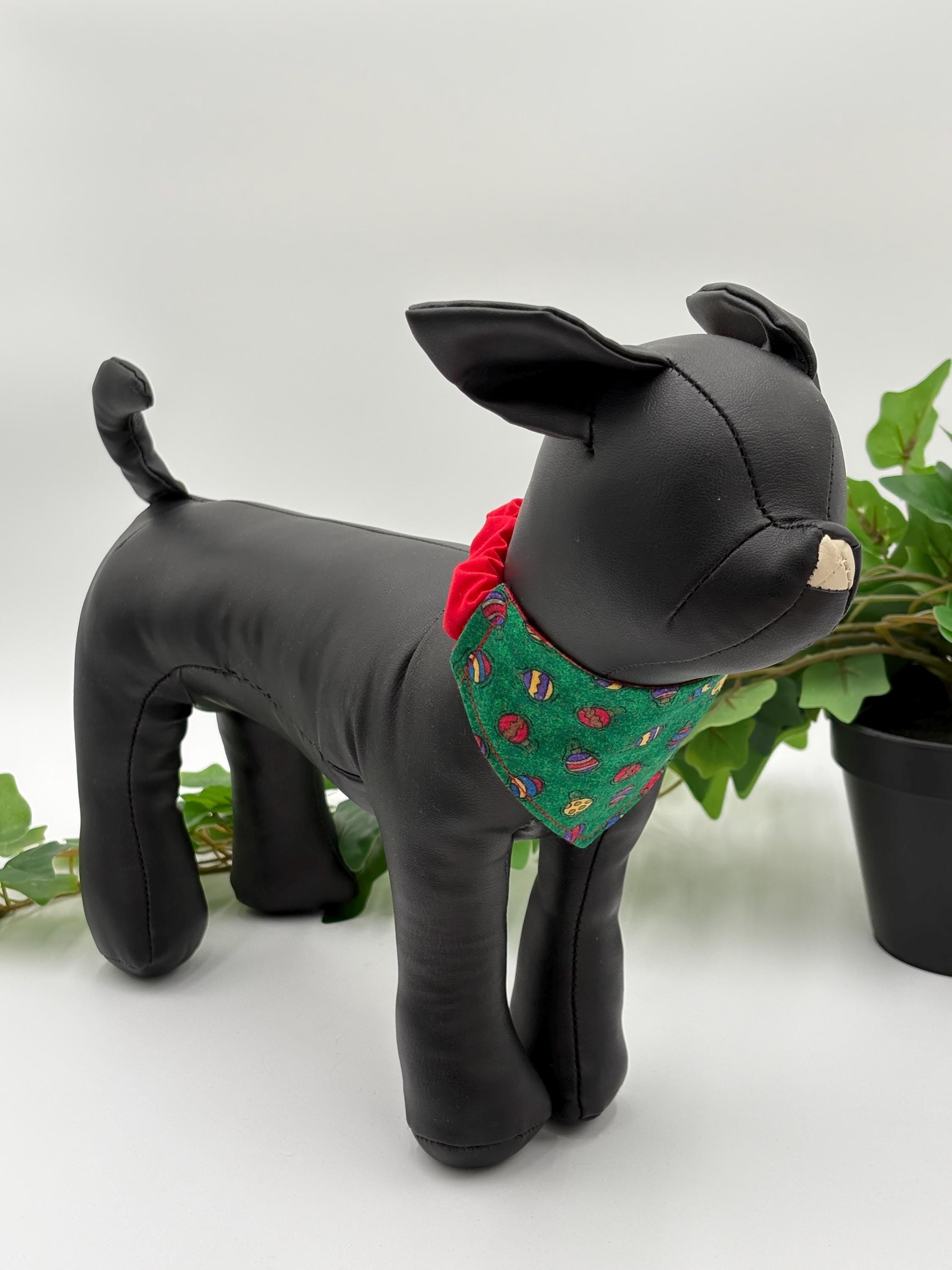 Black dog mannequin with a green bandana featuring mini ornaments on a white background standing next to a potted plant