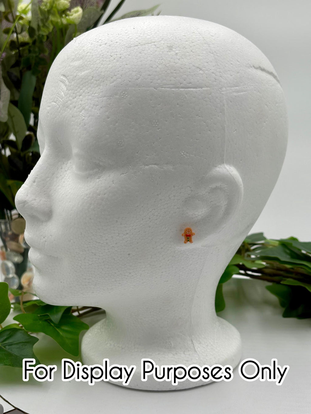 White mannequin head with a small gingerbread man earring on a white surface with green leaves with text "for display purposes only.