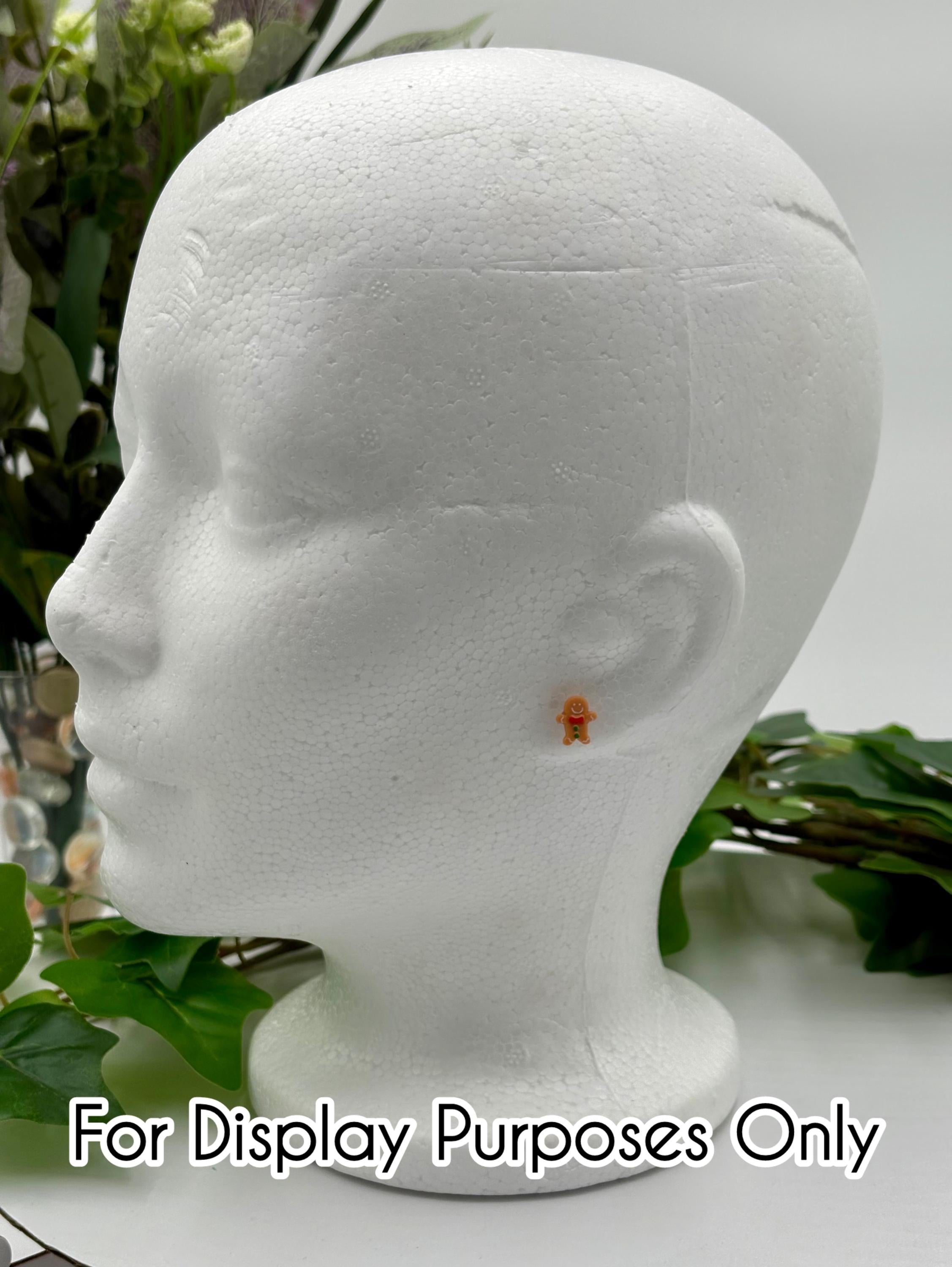 White mannequin head with a small gingerbread man earring on a white surface with green leaves with text "for display purposes only.
