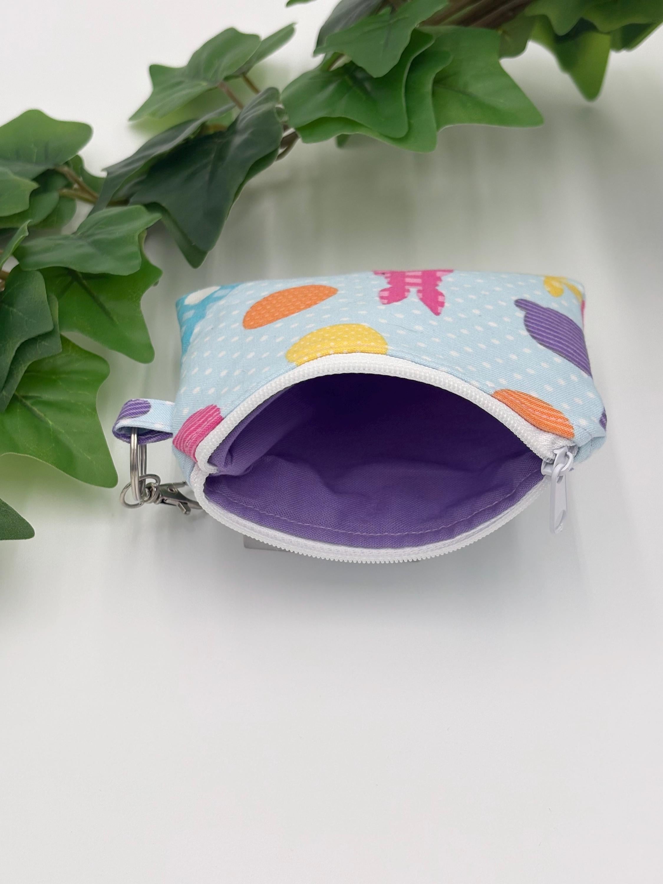 Small coin purse keychain with blue and pastel eggs and bunnies pattern with white zipper opened showing purple lining on a white background with green leaves