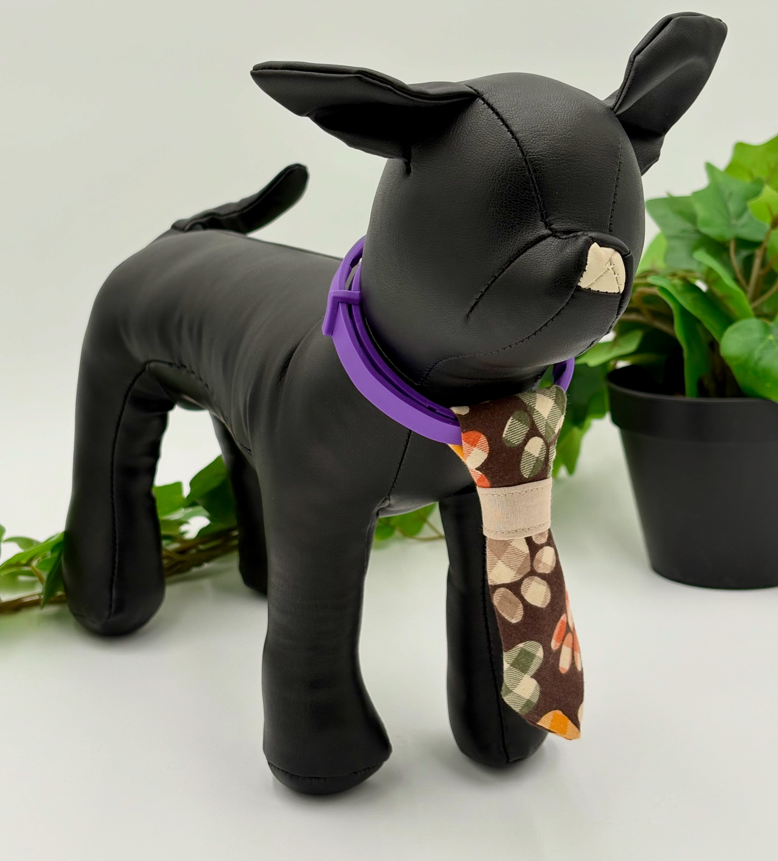 Black dog mannequin with a brown with plaid autumn colored paws necktie on a white background with green potted plant