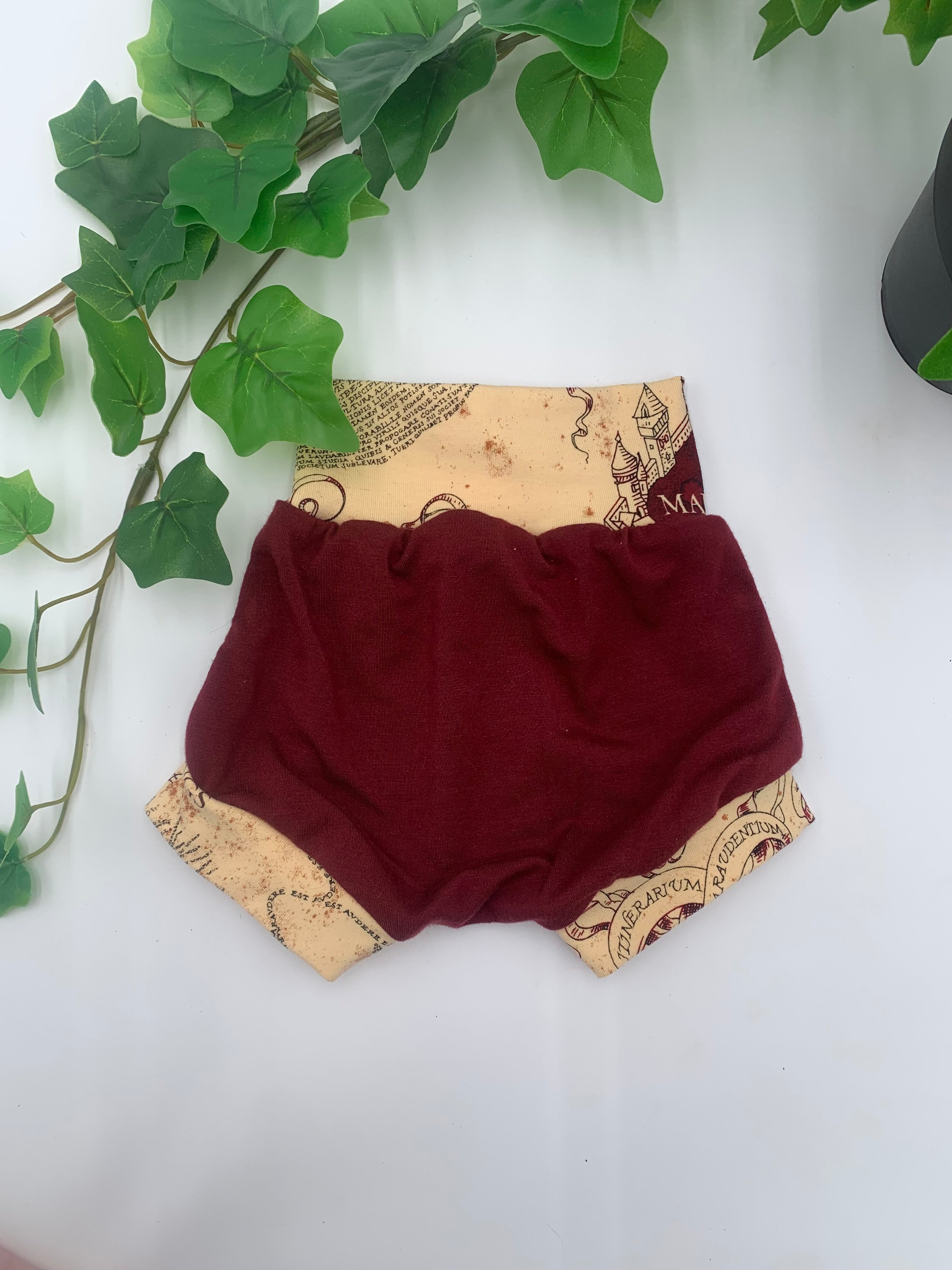 Maroon baby bloomers with beige patterned waistband on a white background with greenery