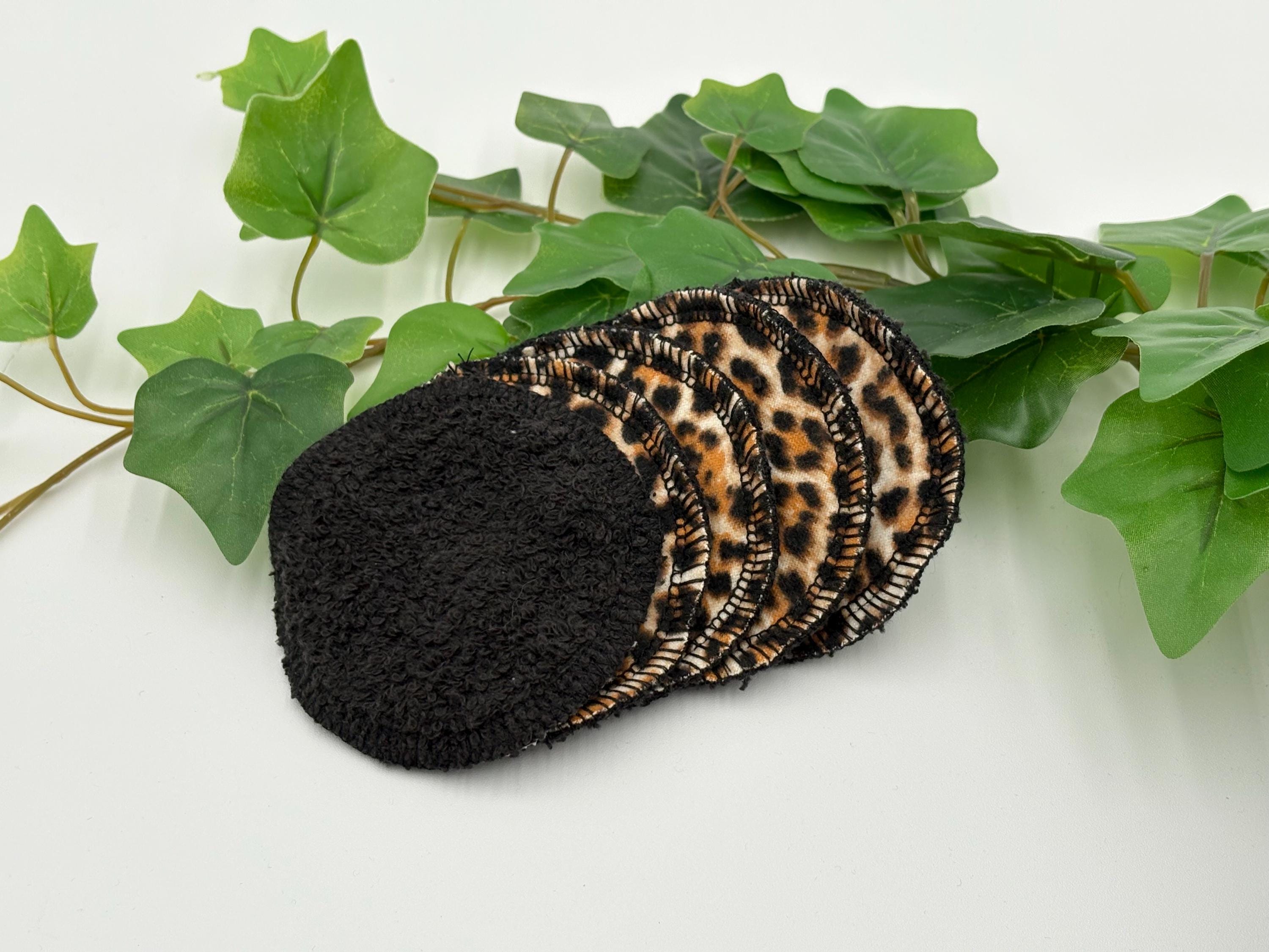Set of five reusable makeup remover pads with leopard print and black fabric, surrounded by green leaves on a white background.