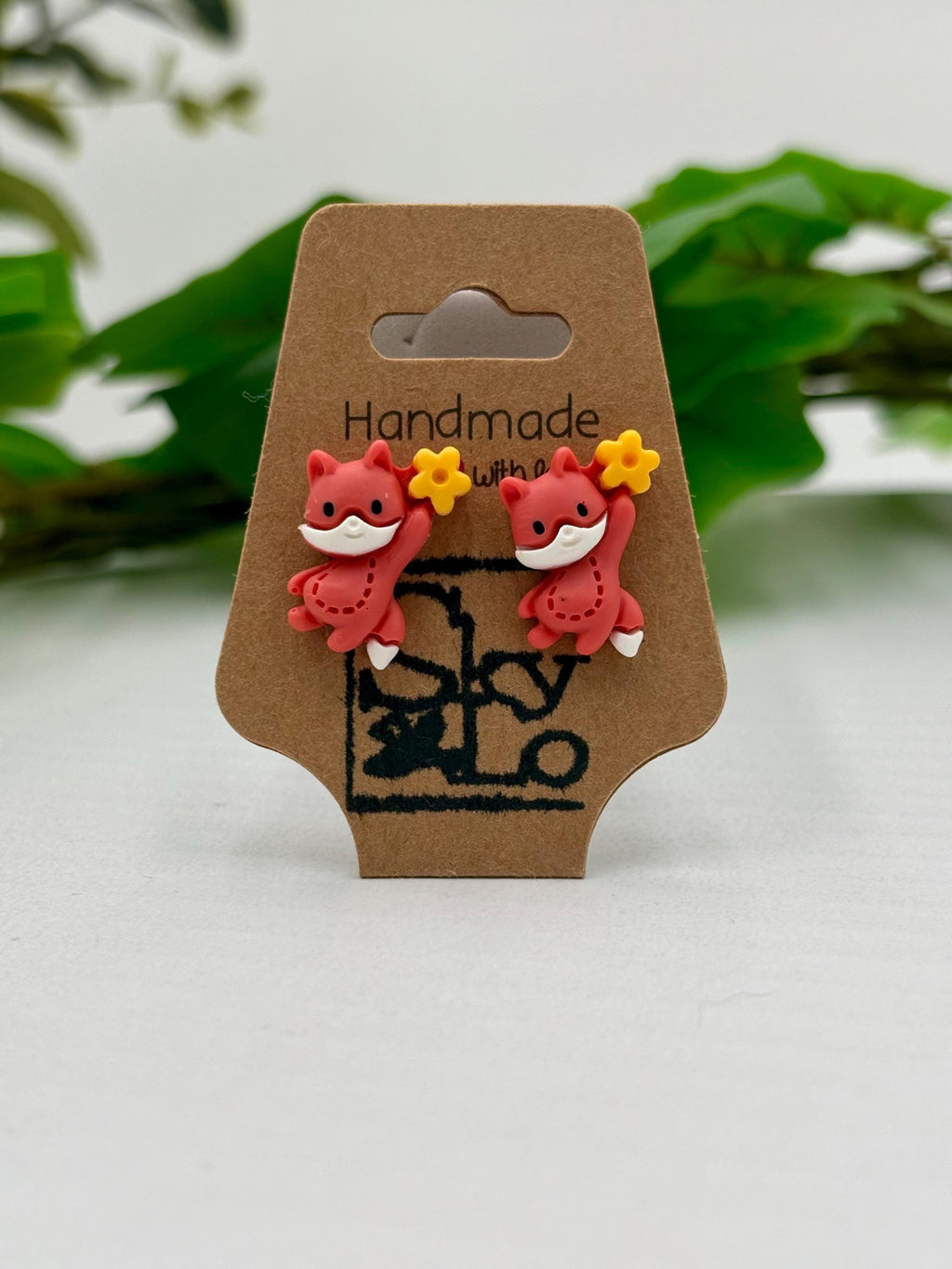 Handmade fox-shaped earrings on a brown card with green leaves in the background