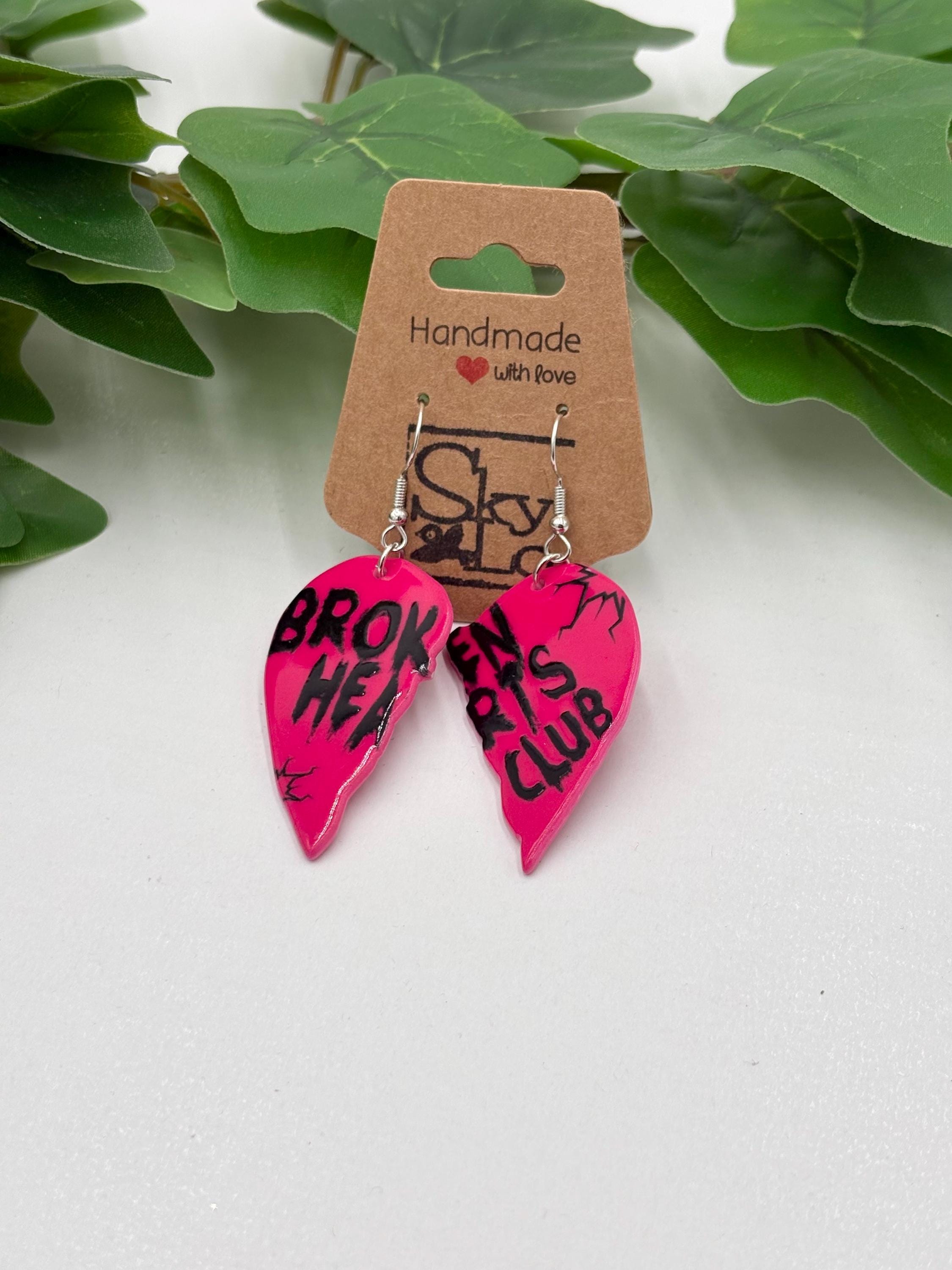 Pink heart-shaped earrings with black text "Broken hearts club" on a brown 'Handmade with Love' card, against a green leafy background.
