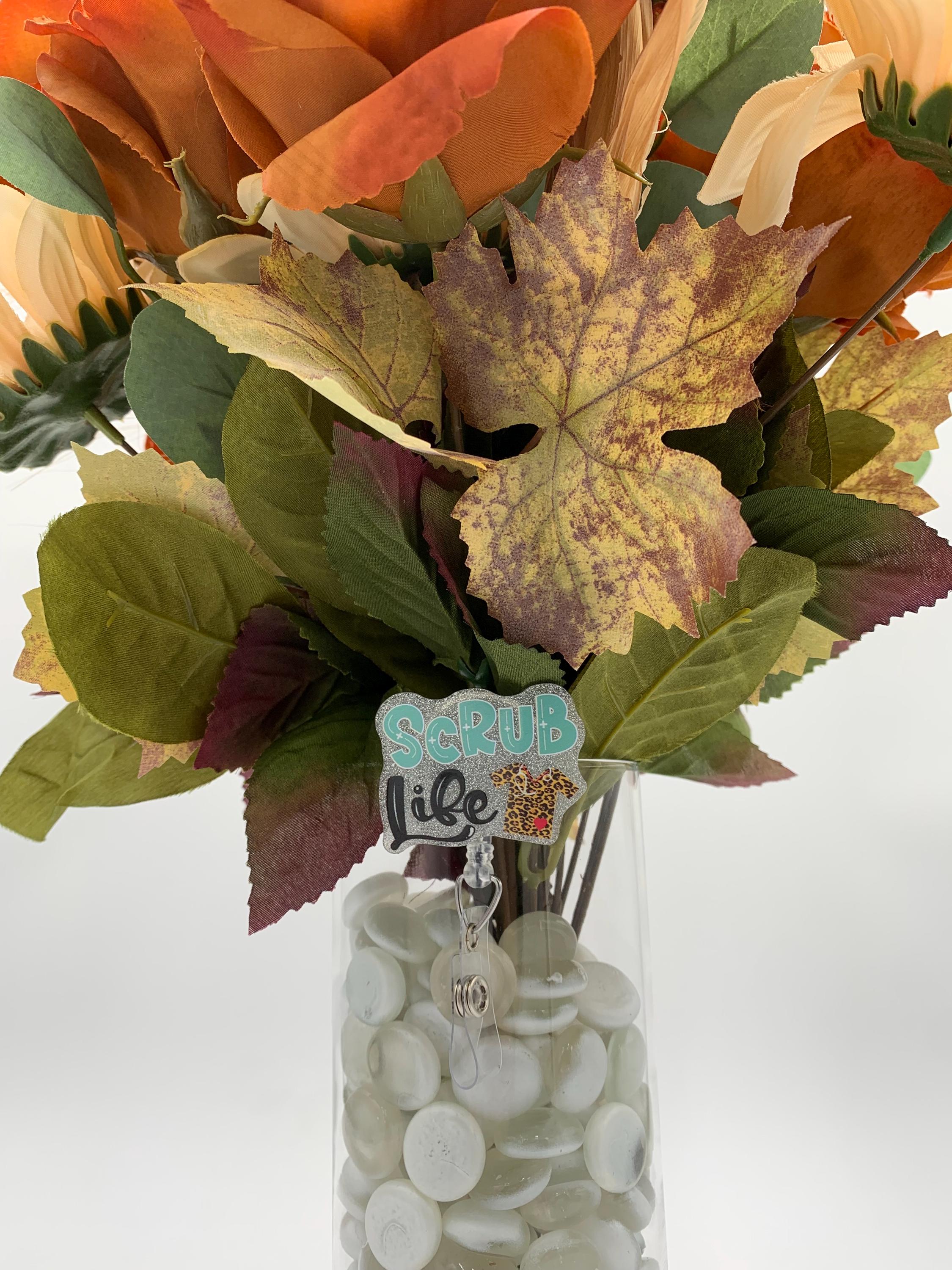 Autumn-themed floral arrangement with leaves and flowers in a clear vase on a white background with a badge reel that says "Scrub Life"