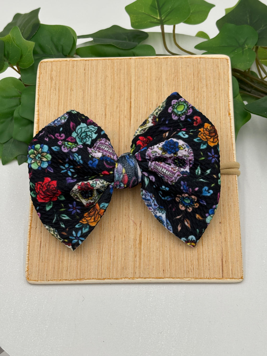 Day of the dead sugar skulls themed big bow nylon headband on a wooden board with a white background and greenery 