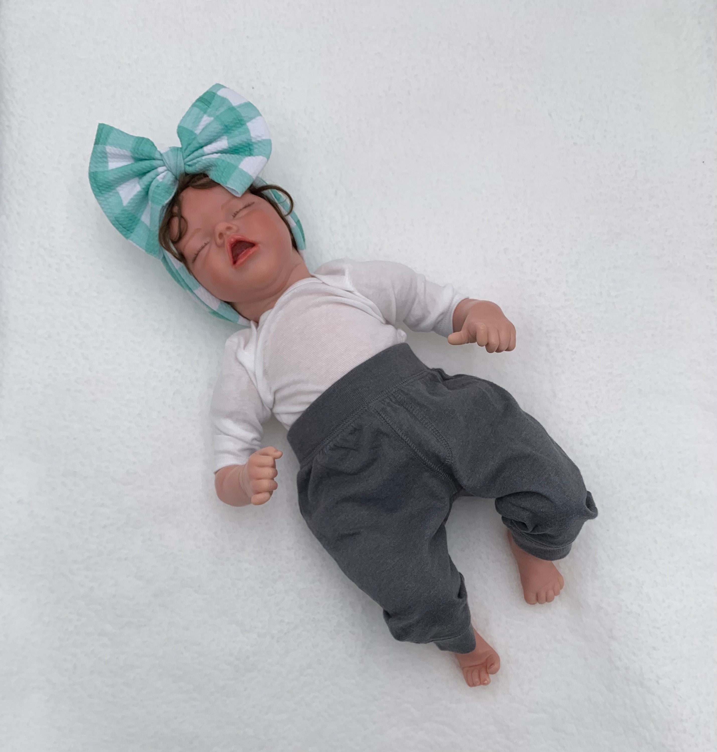Newborn baby doll lying on a white surface wearing a large blue checkered bow headband, white shirt, and gray pants.