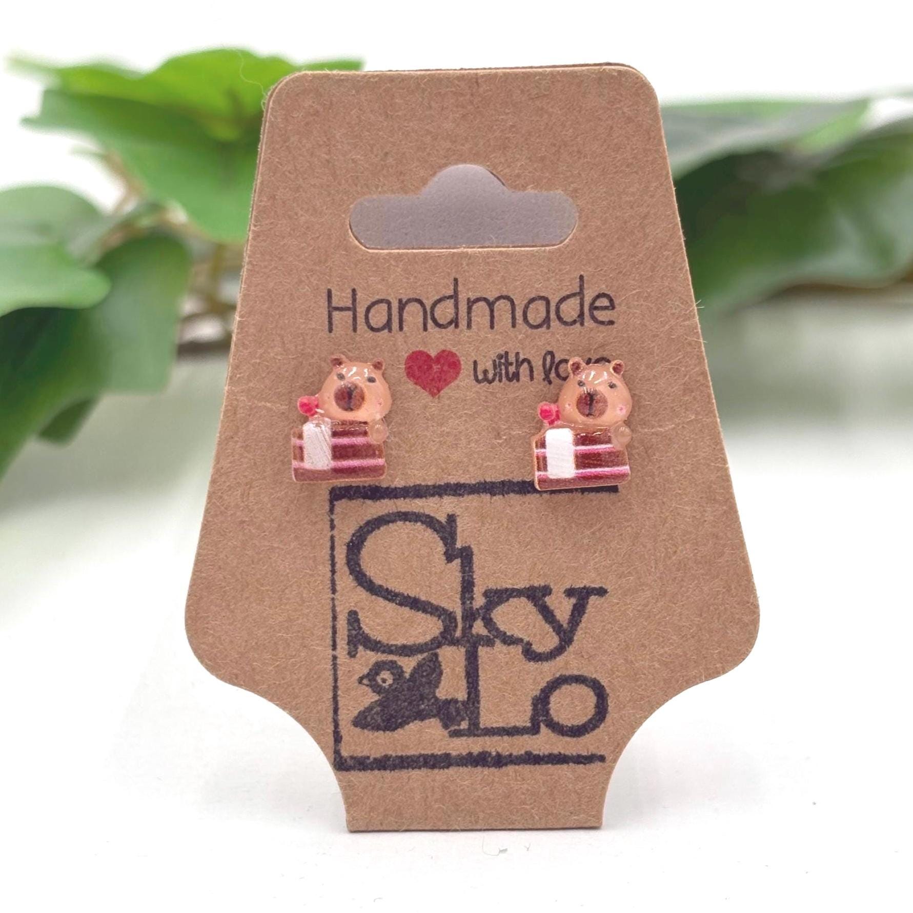 Handmade capybara with chocolate cake earrings on a branded card with a blurred green plant background