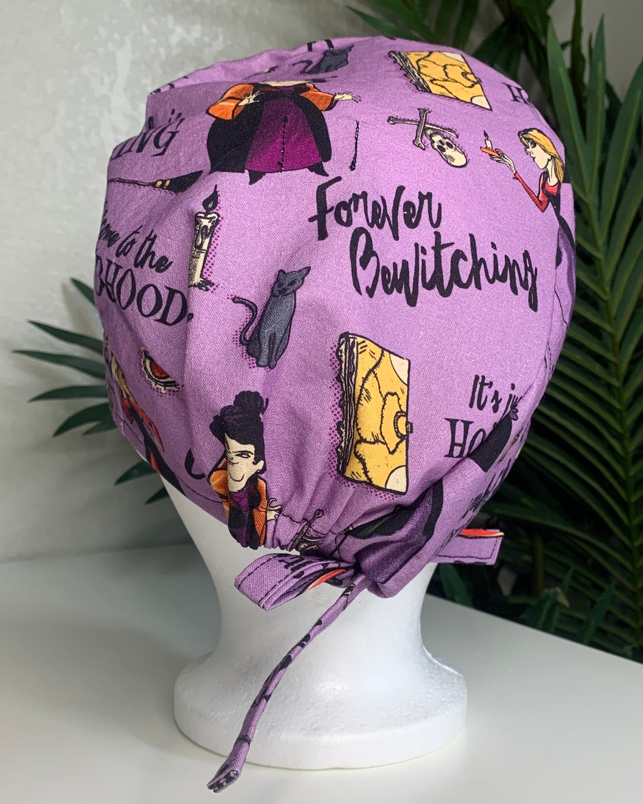 Purple Hocus Pocus patterned scrub cap on a mannequin head with a white background with plant