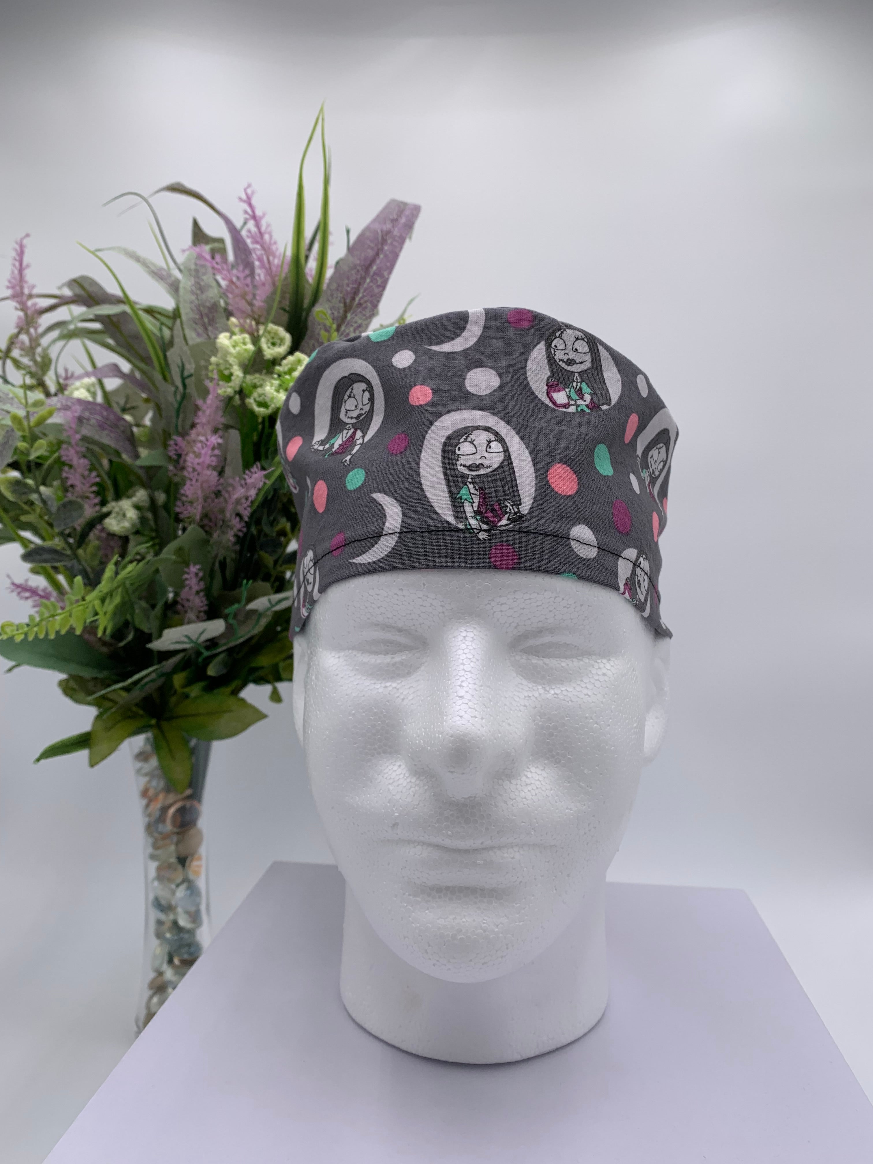Gray polka dot nightmare before Christmas scrub cap on a mannequin head with flowers, against a white background