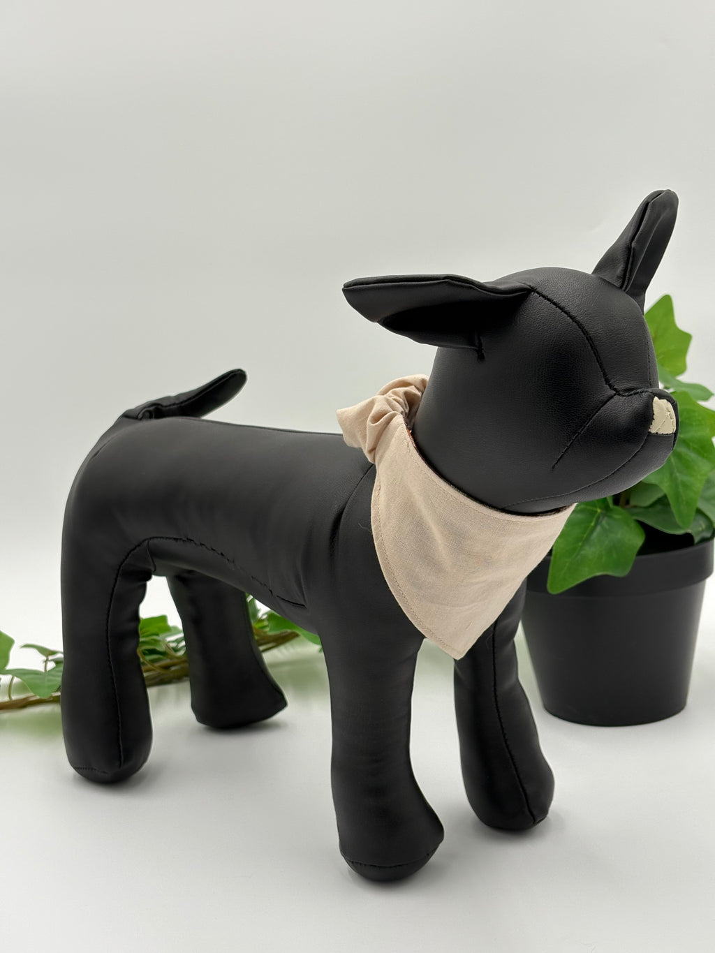 Black dog mannequin wearing a khaki scrunchie bandana on a white background with green leaves.