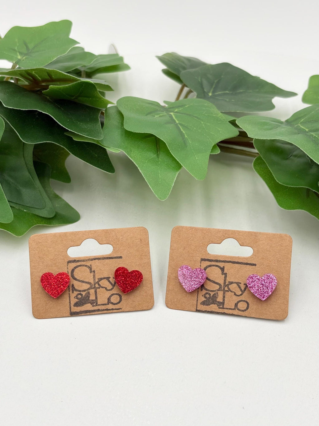 Two pairs of sparkling heart-shaped earrings on cardboard cards with a green leafy background.