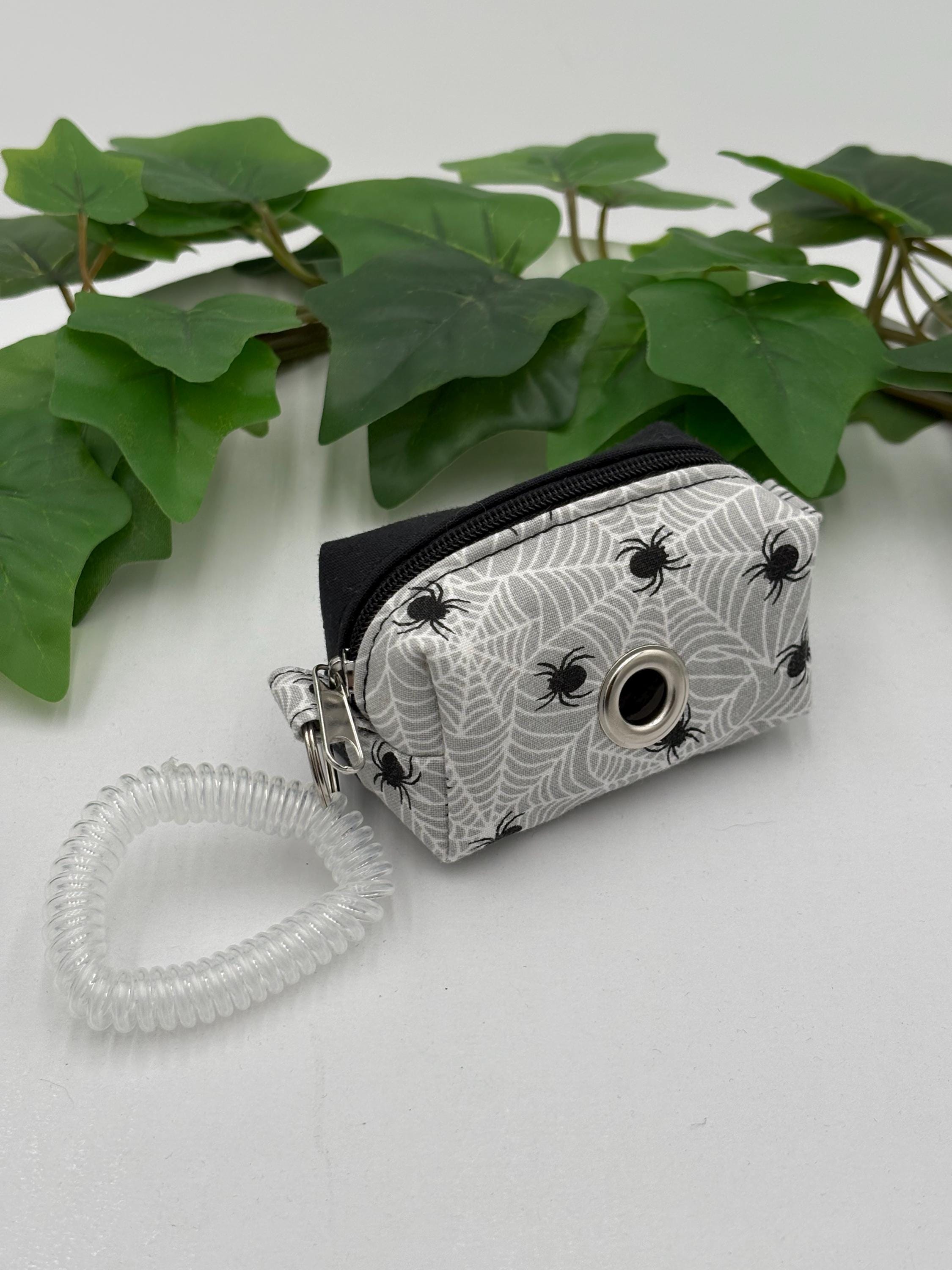 Spider web patterned dog poop bag holder with clear coiled strap on a light background with green leaves.