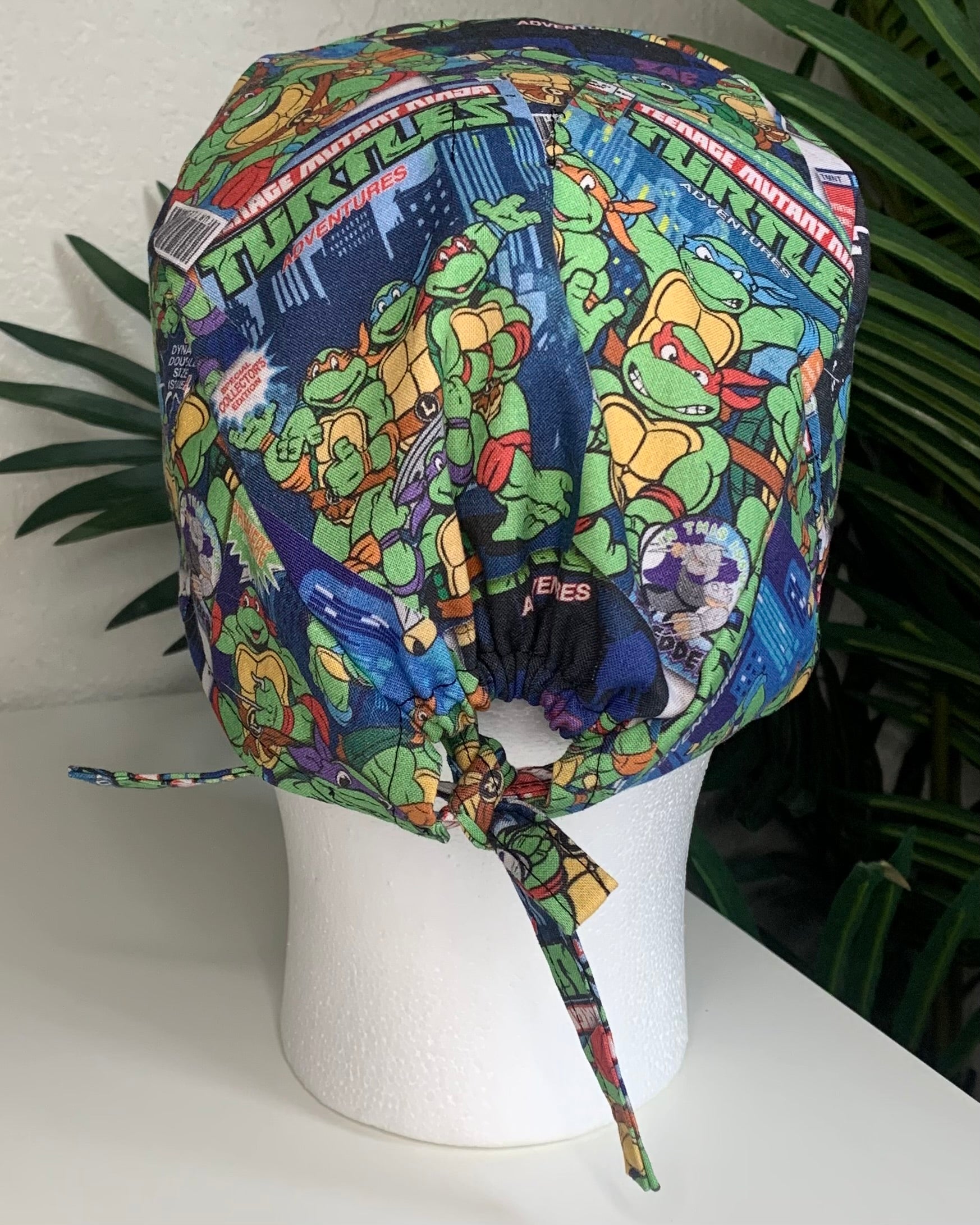 Colorful Teenage Mutant Ninja’ Turtles scrub cap on a mannequin head with a plant, against a white background