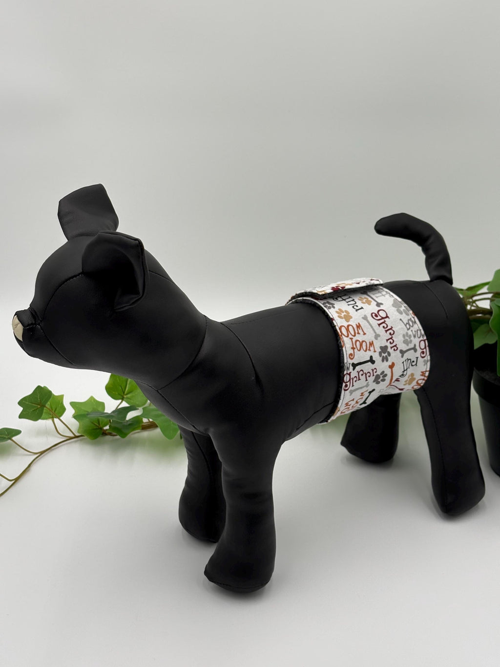 Black dog mannequin with a patterned belly band on a white background standing next to potted plant