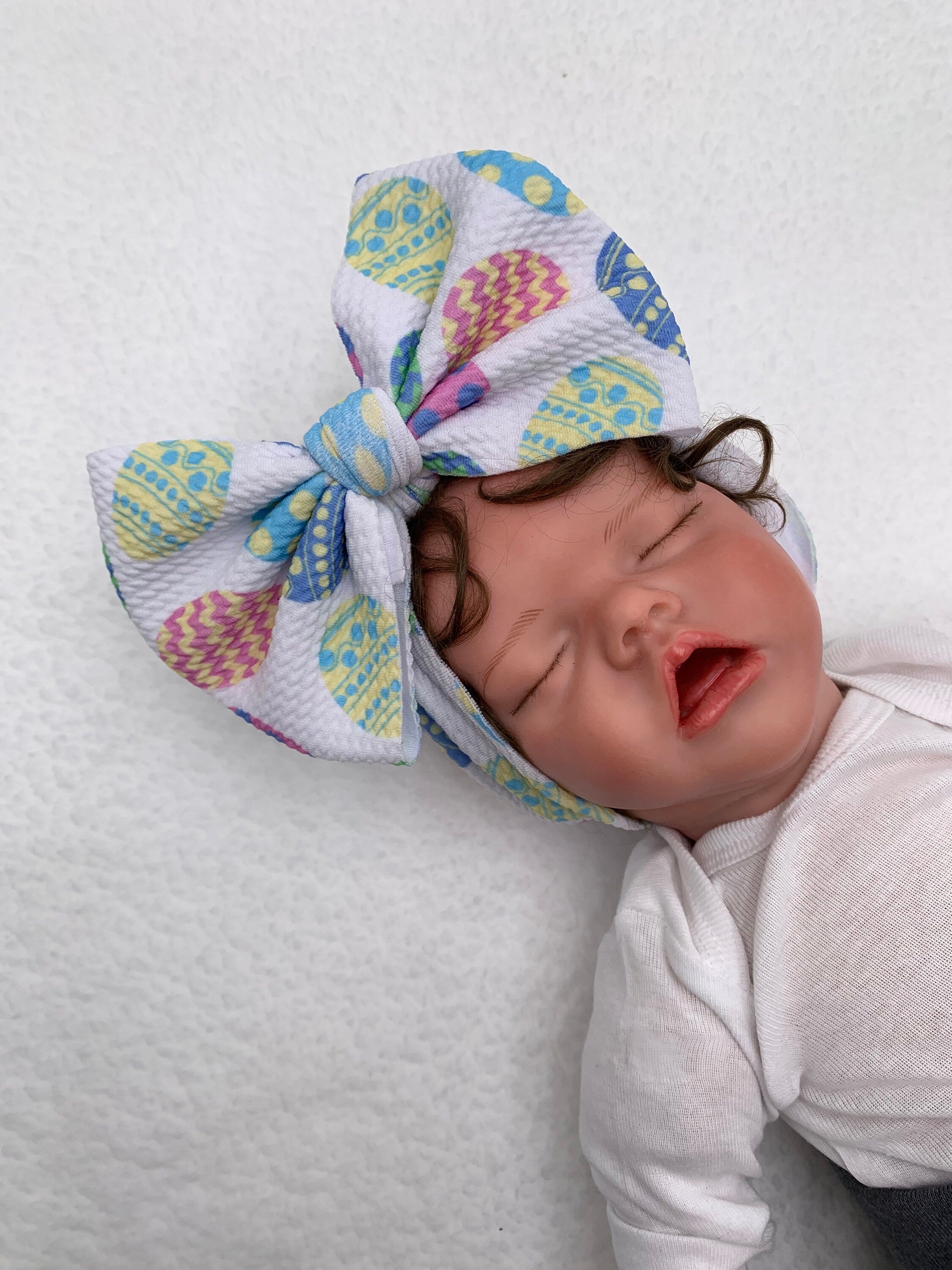 Newborn baby doll wearing a White Headband with a decorative easter eggs themed big bow on a mannequin head silhouette against a white background