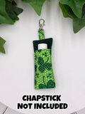 Green shamrock-patterned chapstick holder on a white surface with green leaves in the background with text "chapstick not included"