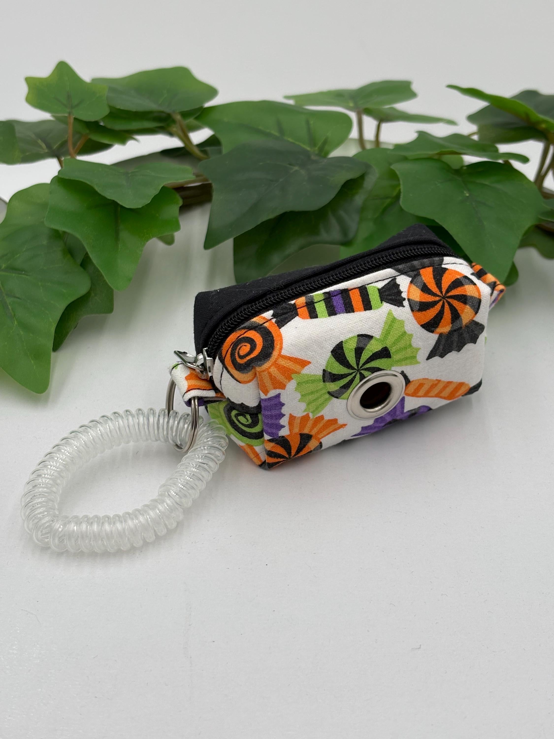 Colorful Halloween-themed dog poop bag holder with a coiled keychain on a white surface with green leaves in the background.