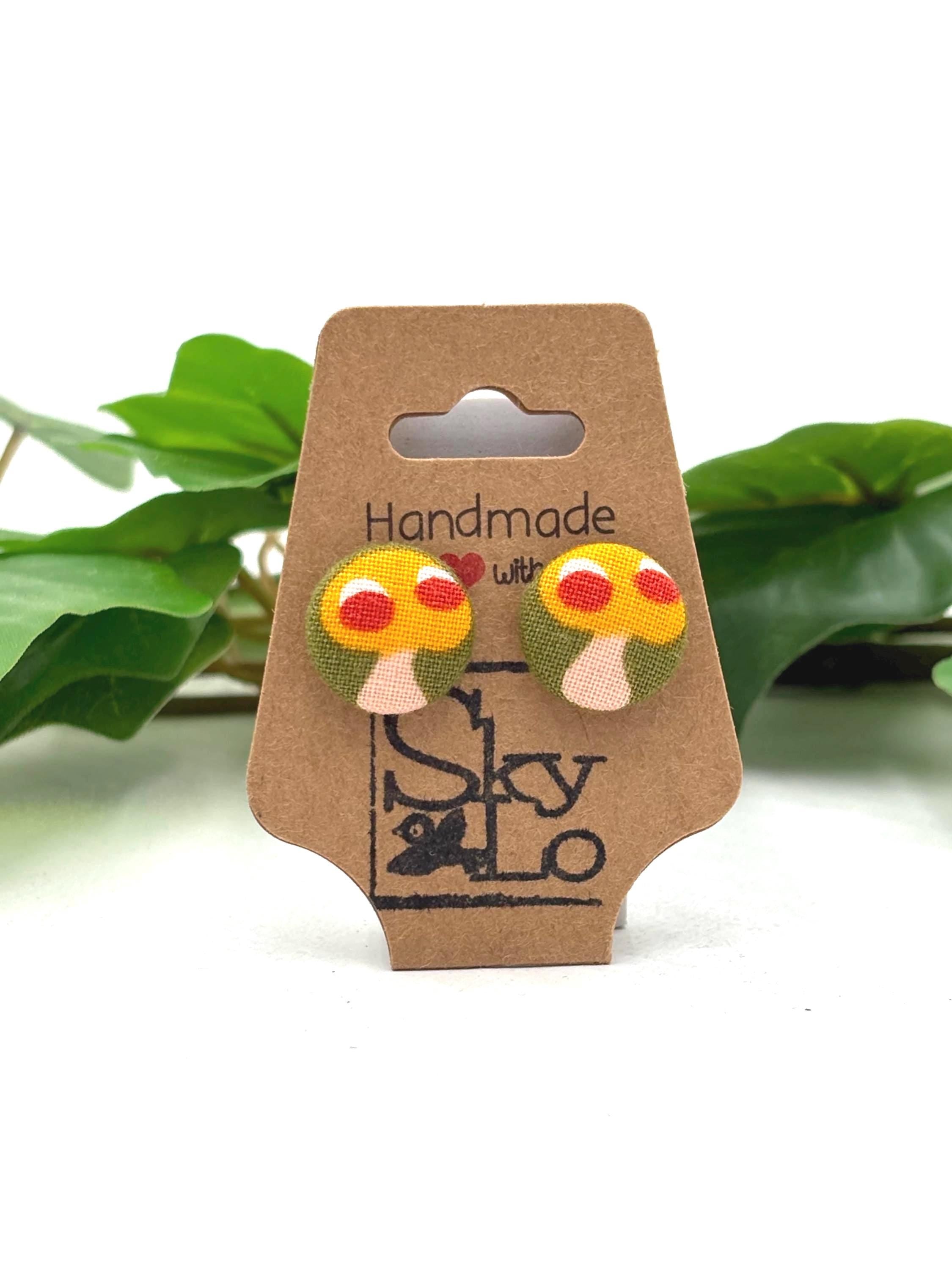 Cardboard earring display card with yellow mushroom  with red dots earrings and 'Handmade with love' and 'SkyLo' branding, against a white background with green leaves.