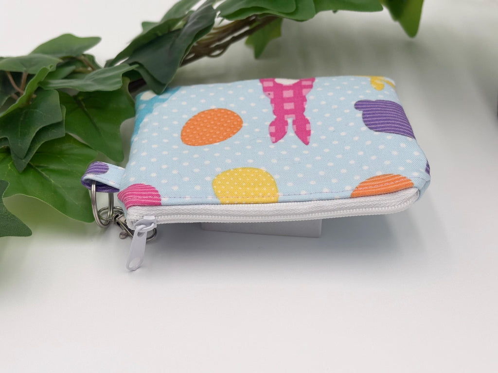 Small coin purse keychain with blue and pastel eggs and bunnies pattern with a white zipper on a white background with green leaves
