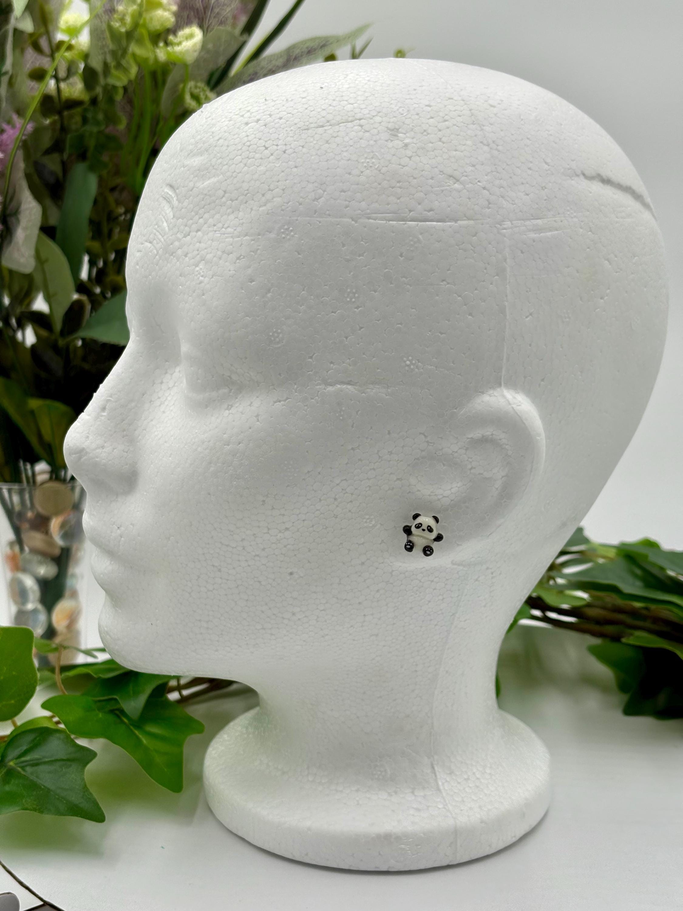 White mannequin head with a small panda shaped earring on a white background with greenery.