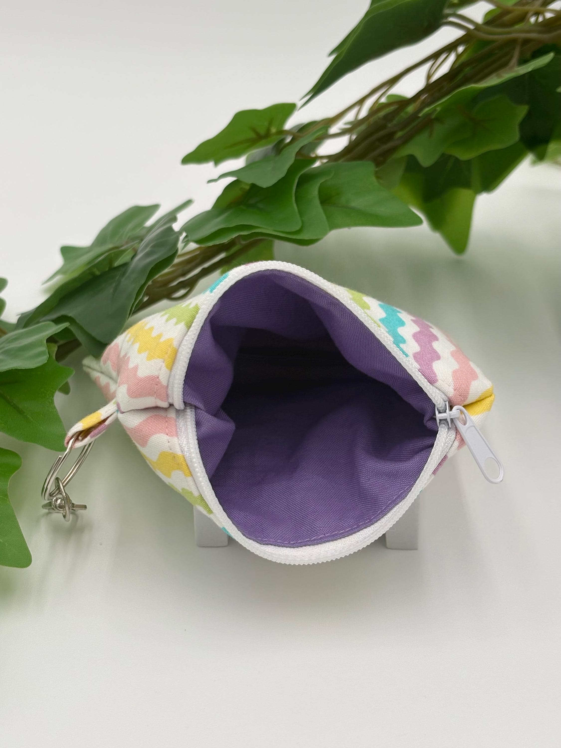 Small coin purse keychain with pastel zigzag pattern with white zipper open showing purple lining on a white background with green leaves 
