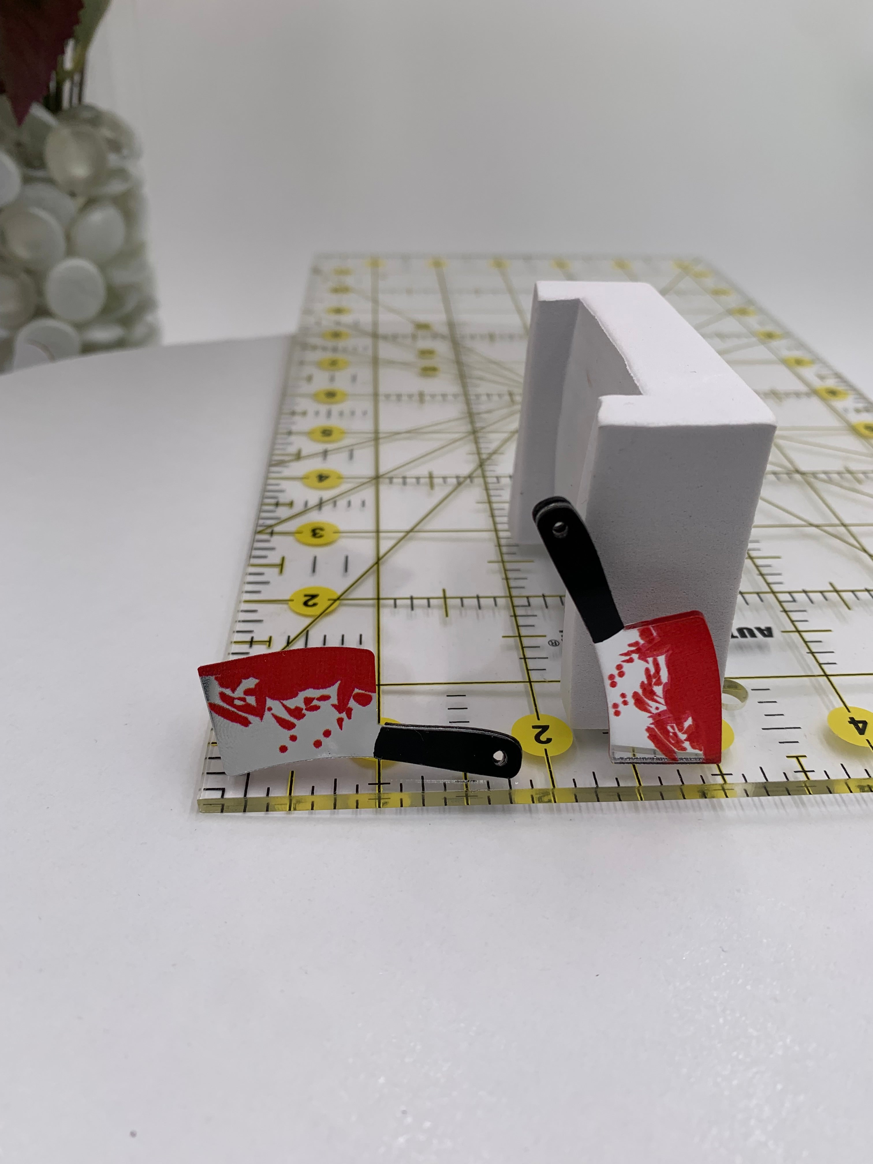 Bloody meat cleaver earring next to a ruler with measurement markings at 2 inches and 1 inch