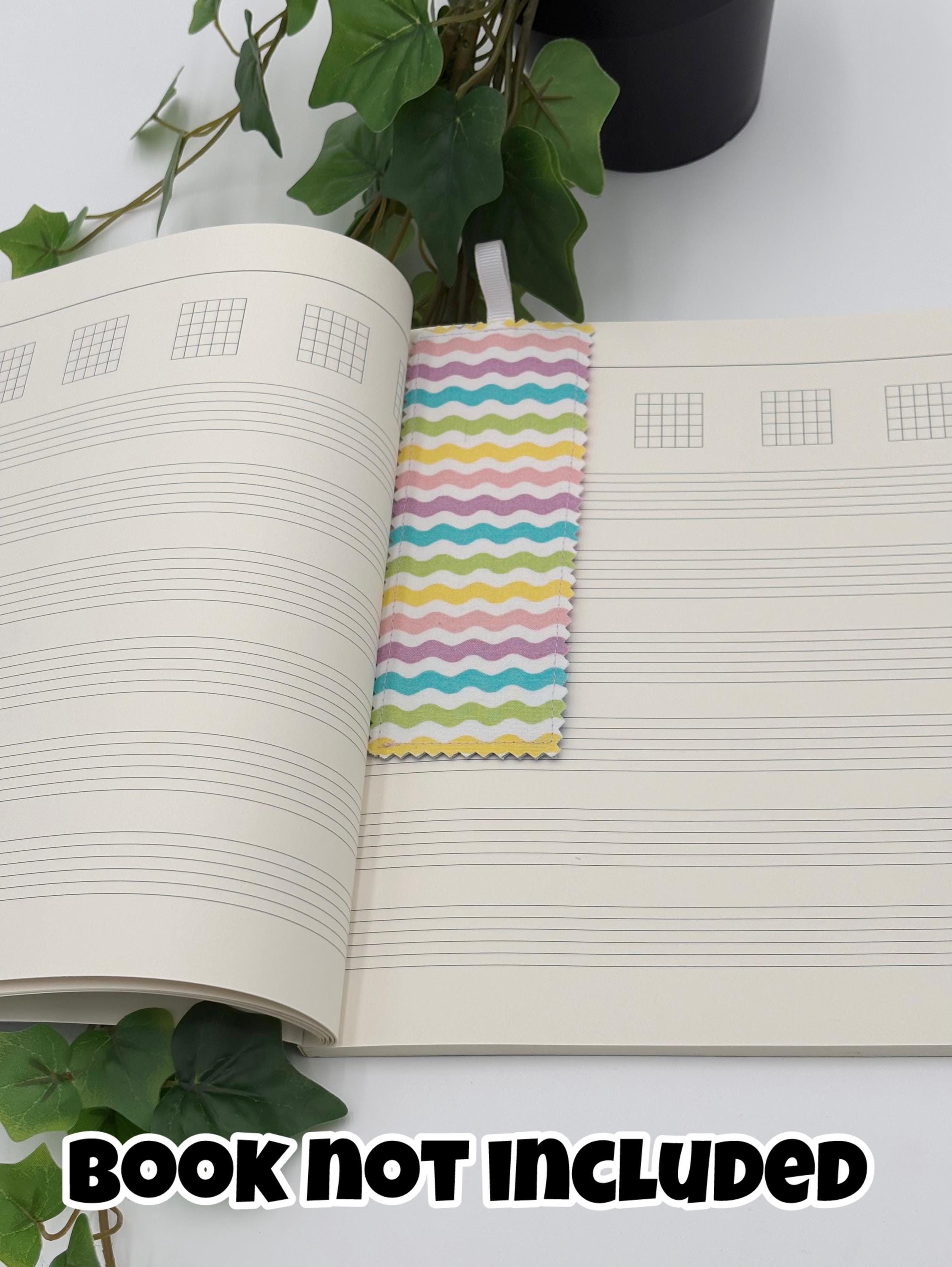 Open notebook with a colorful pastel wavy bookmark on a white surface with green leaves. with text that says "book not included"