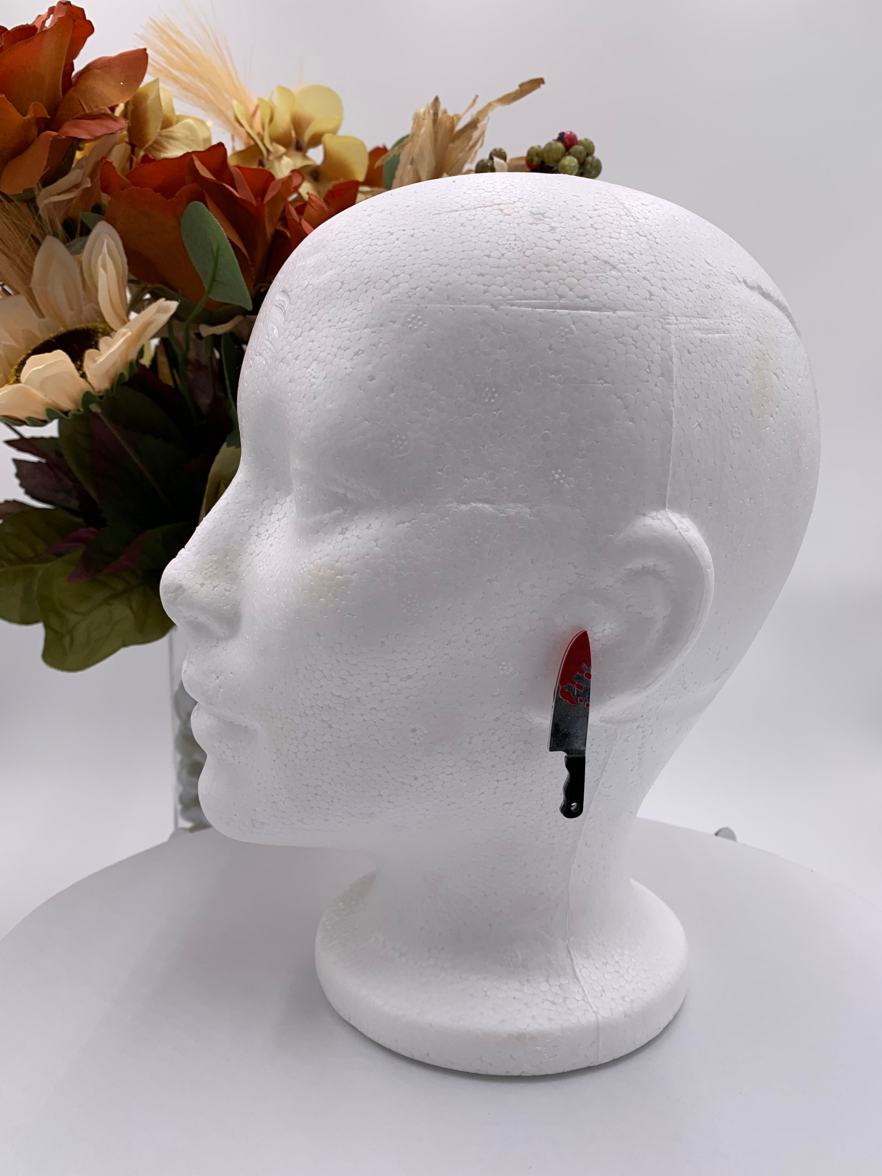 White Mannequin head with bloody butcher knife earrings on a white background with a bouquet of flowers. 