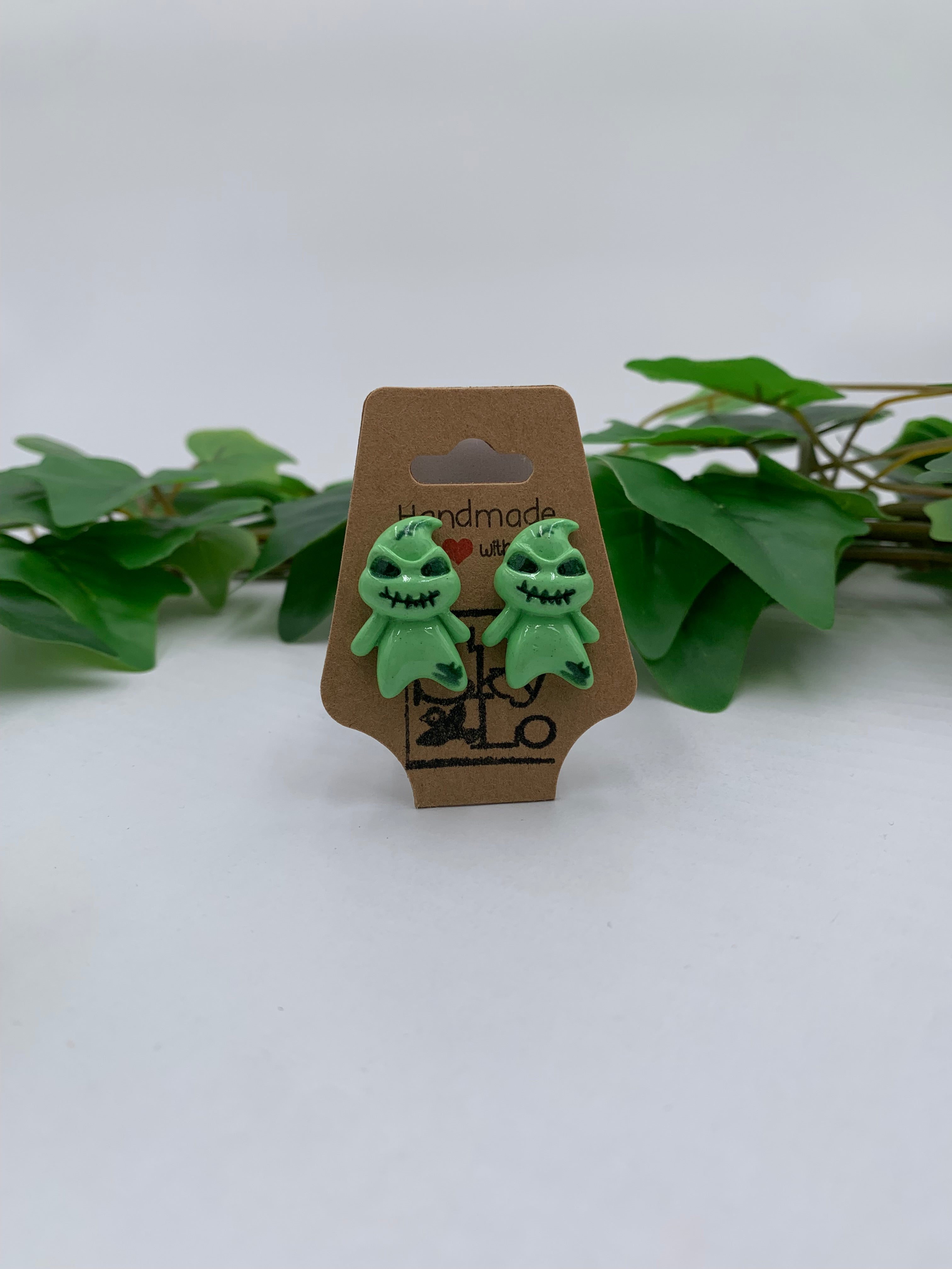 Green boogyman earrings on a cardboard backing with a plant on a white background