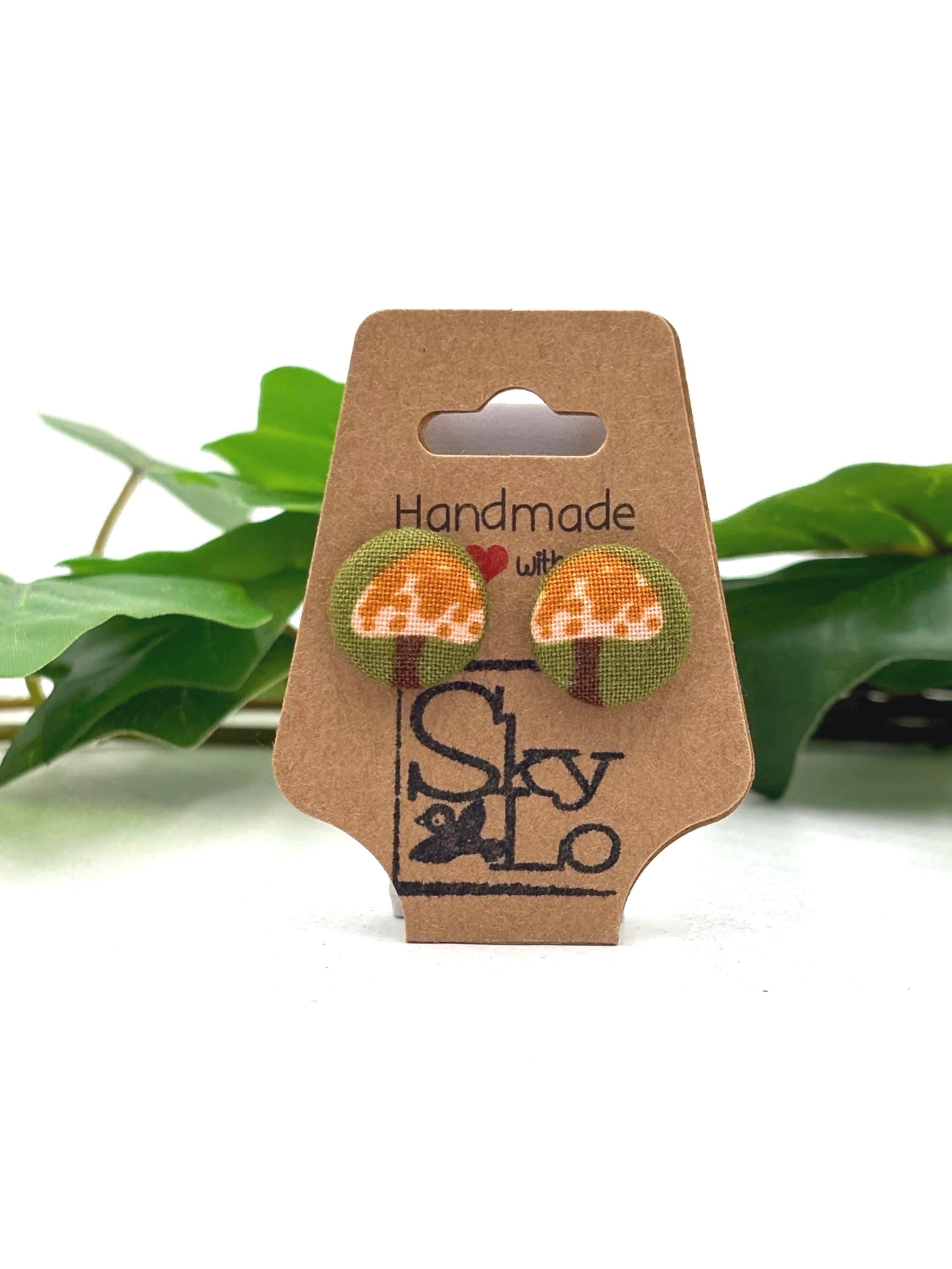 Cardboard earring display card with beige mushroom with brown dots earrings and 'Handmade with love' and 'SkyLo' branding, against a white background with green leaves.