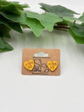 Yellow heart-shaped earrings with 'Kiss Me' text on a cardboard backing with 'SkyLo' branding, placed on a light surface with green leaves in the background.